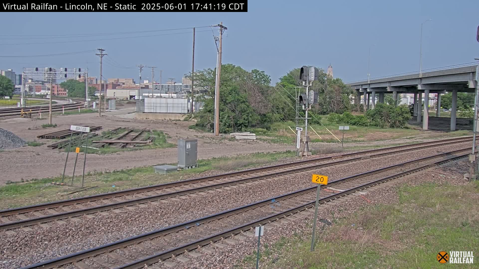 A sunny day reveals railroad tracks stretching toward a highway overpass and a cityscape in the distance, with some construction materials visible near the tracks.
