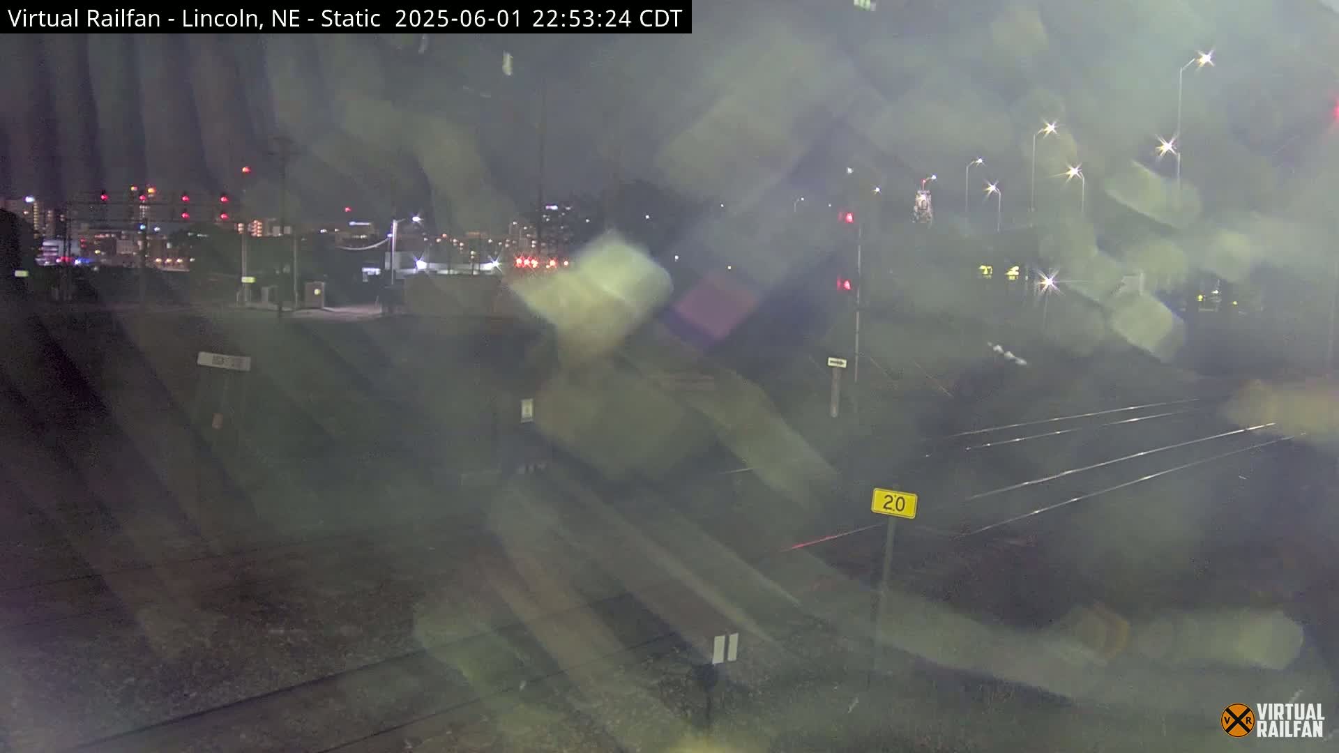 A nighttime view of railroad tracks and a road intersection, with distant city lights visible under hazy or foggy conditions.