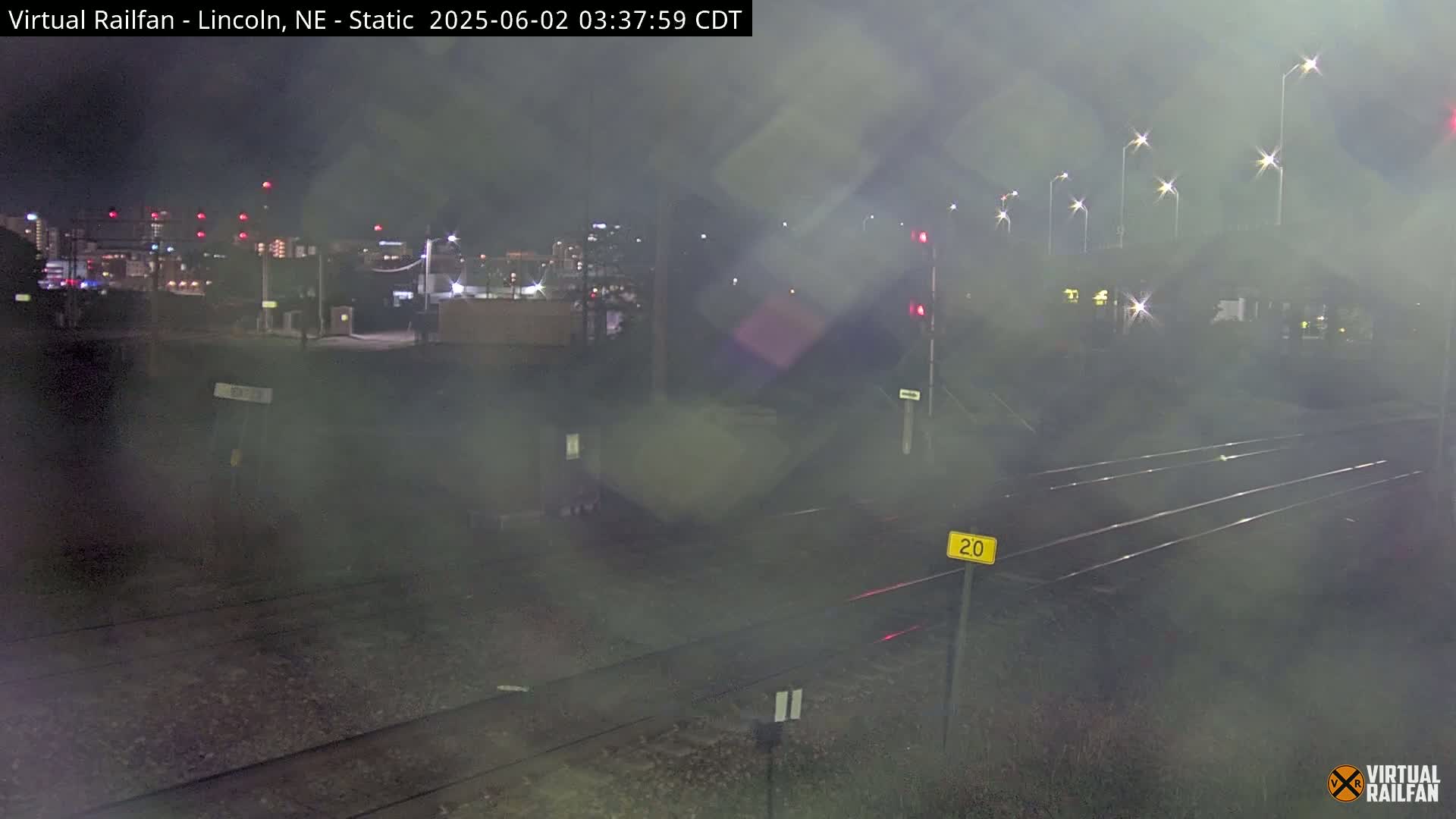 A nighttime view of a hazy, dimly lit train track crossing, with a city skyline in the background.