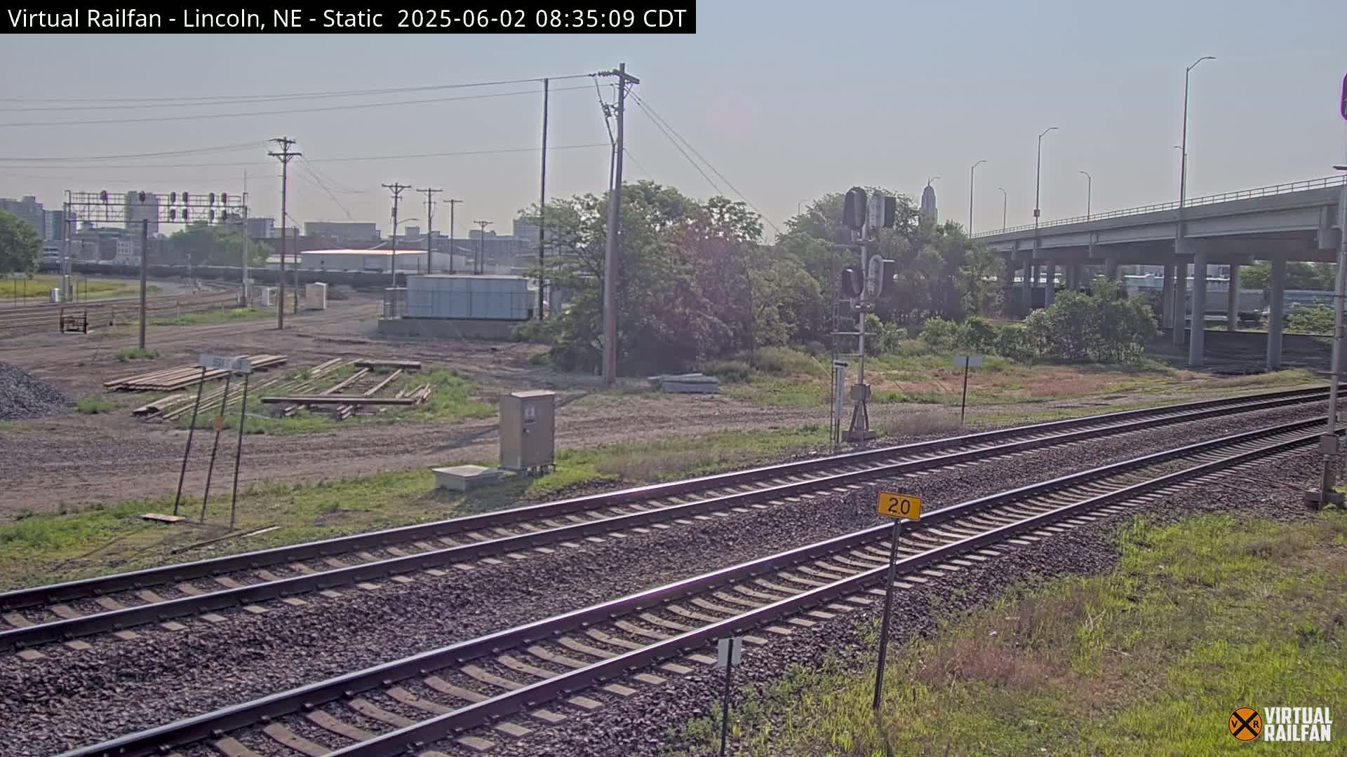 A view of railroad tracks under a mostly clear sky, with some vegetation and industrial buildings in the background.