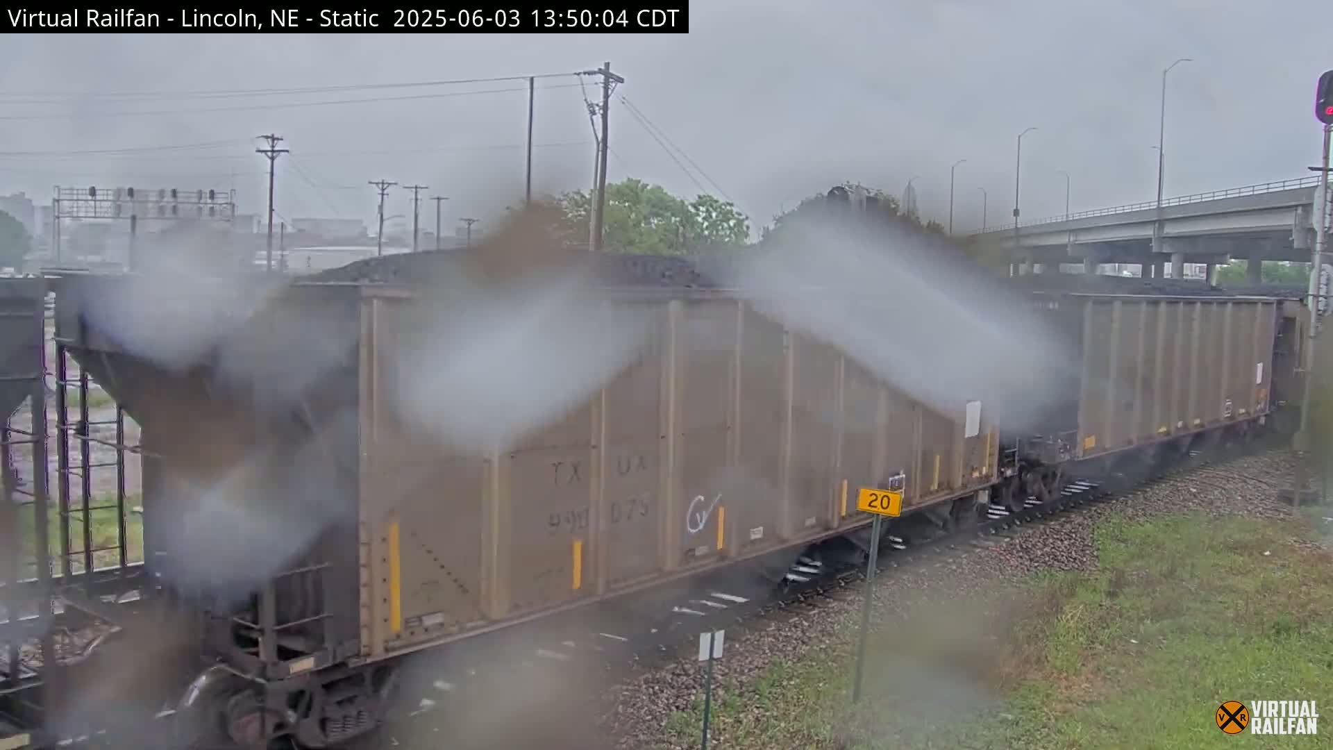 A long freight train carrying coal is seen on a rainy day, traveling along railroad tracks that pass under a highway overpass.