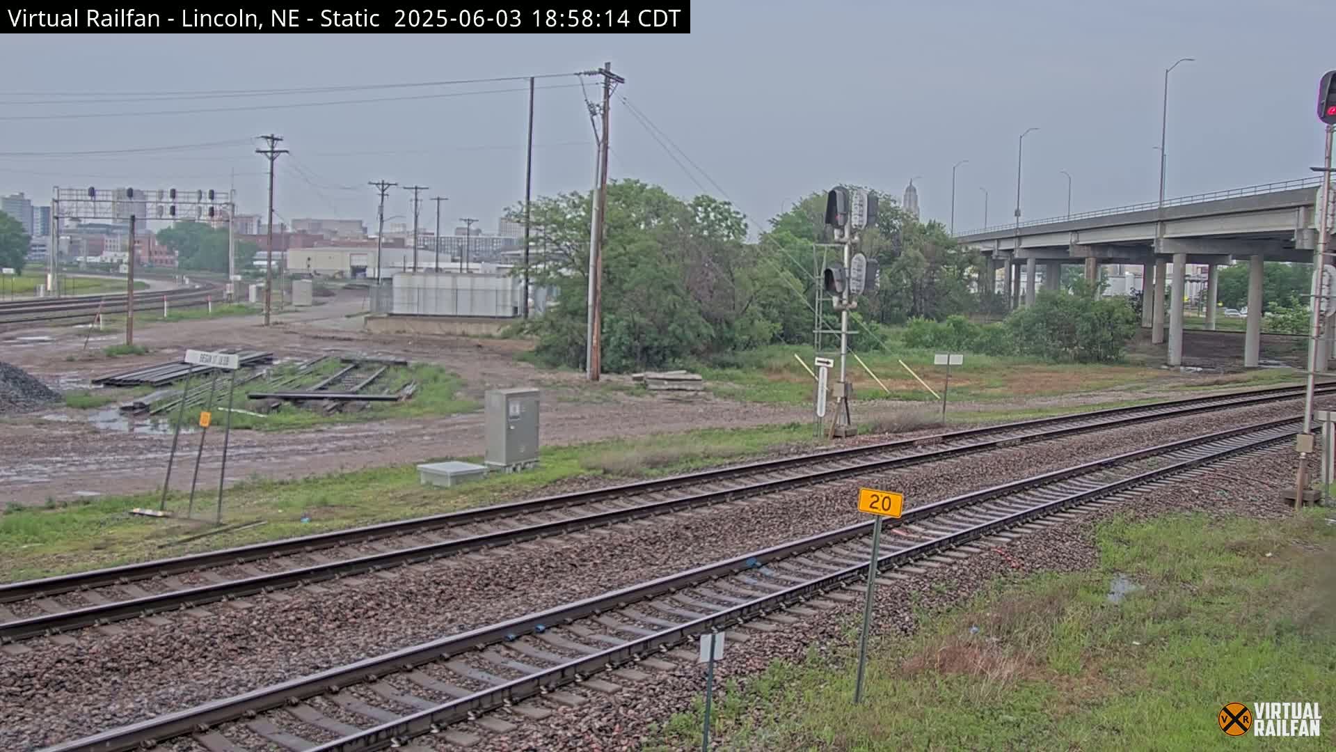 A view of railroad tracks under an overcast sky, with a highway overpass in the background and some industrial buildings visible in the distance.
