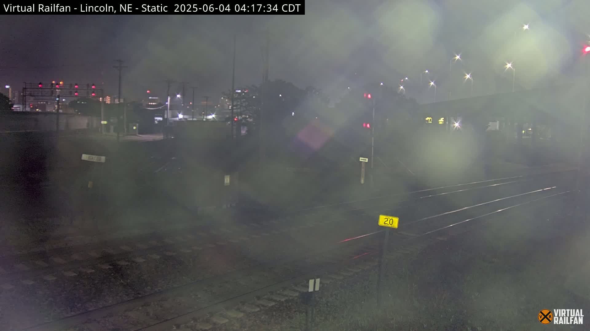 A nighttime view of railroad tracks and a crossing, shrouded in fog or mist, with distant city lights visible.