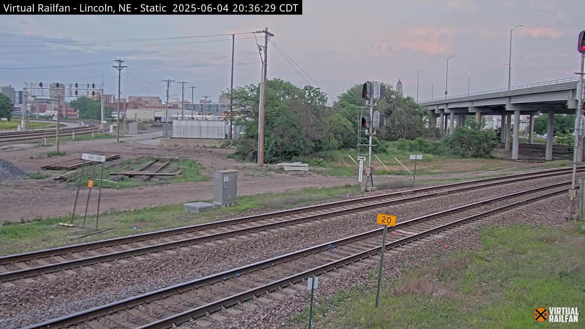 Railroad tracks extend toward a highway overpass under a twilight sky, with a cityscape visible in the distance.