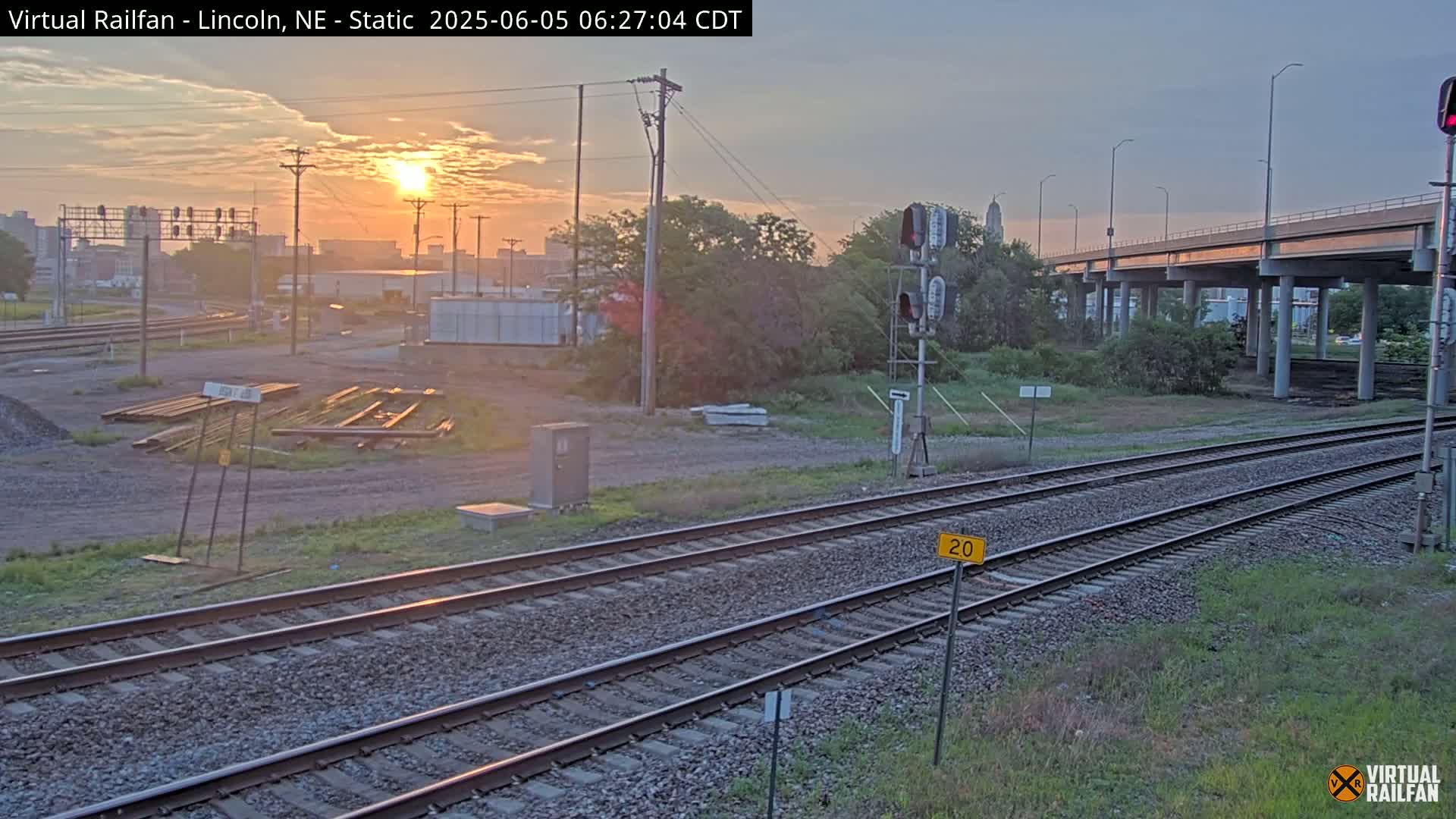 A sunrise illuminates a railway scene with two sets of tracks near a highway overpass, some construction materials, and vegetation.