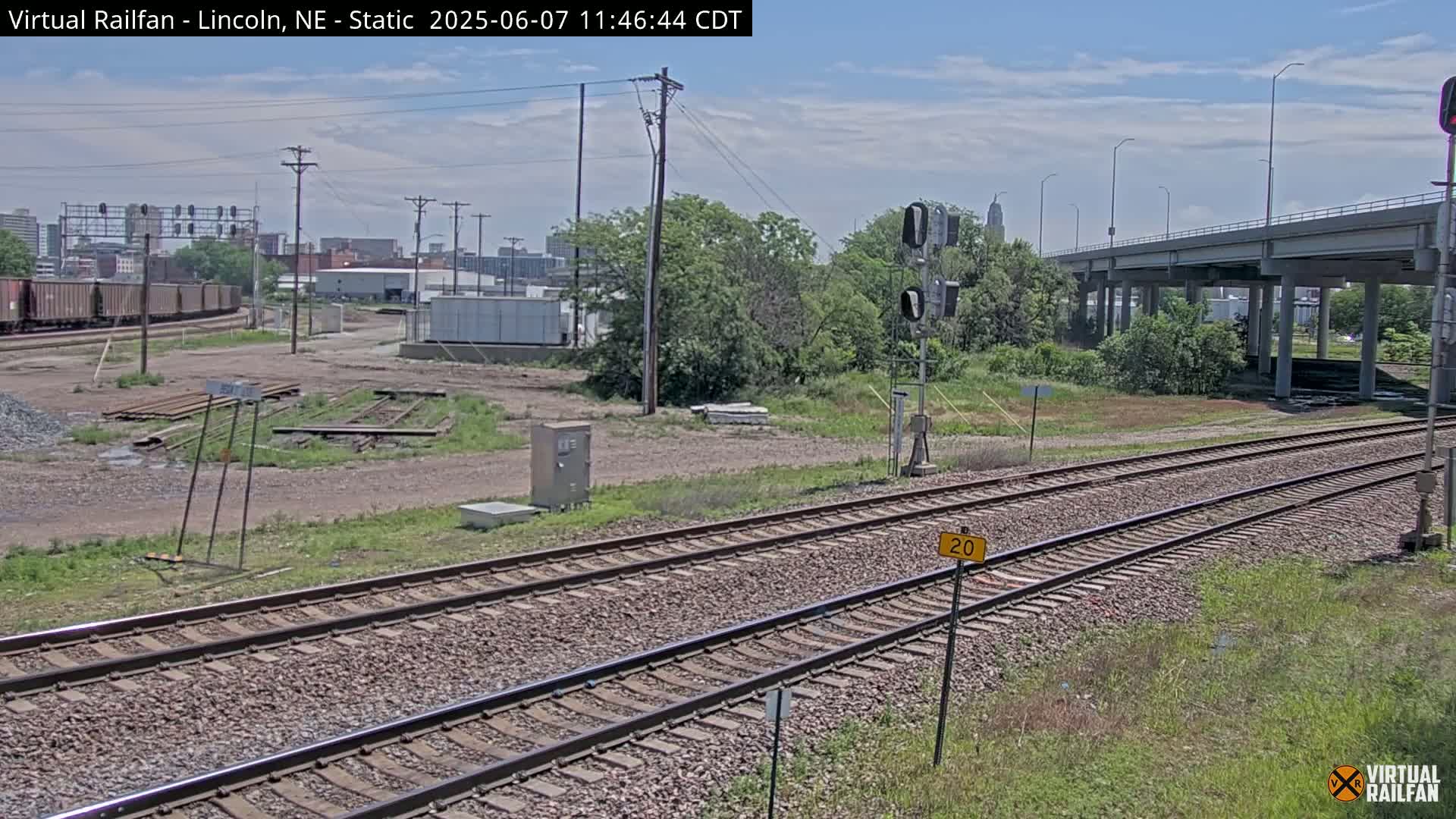 Under a partly cloudy sky, a view of train tracks stretches toward a highway overpass and a cityscape in the distance.