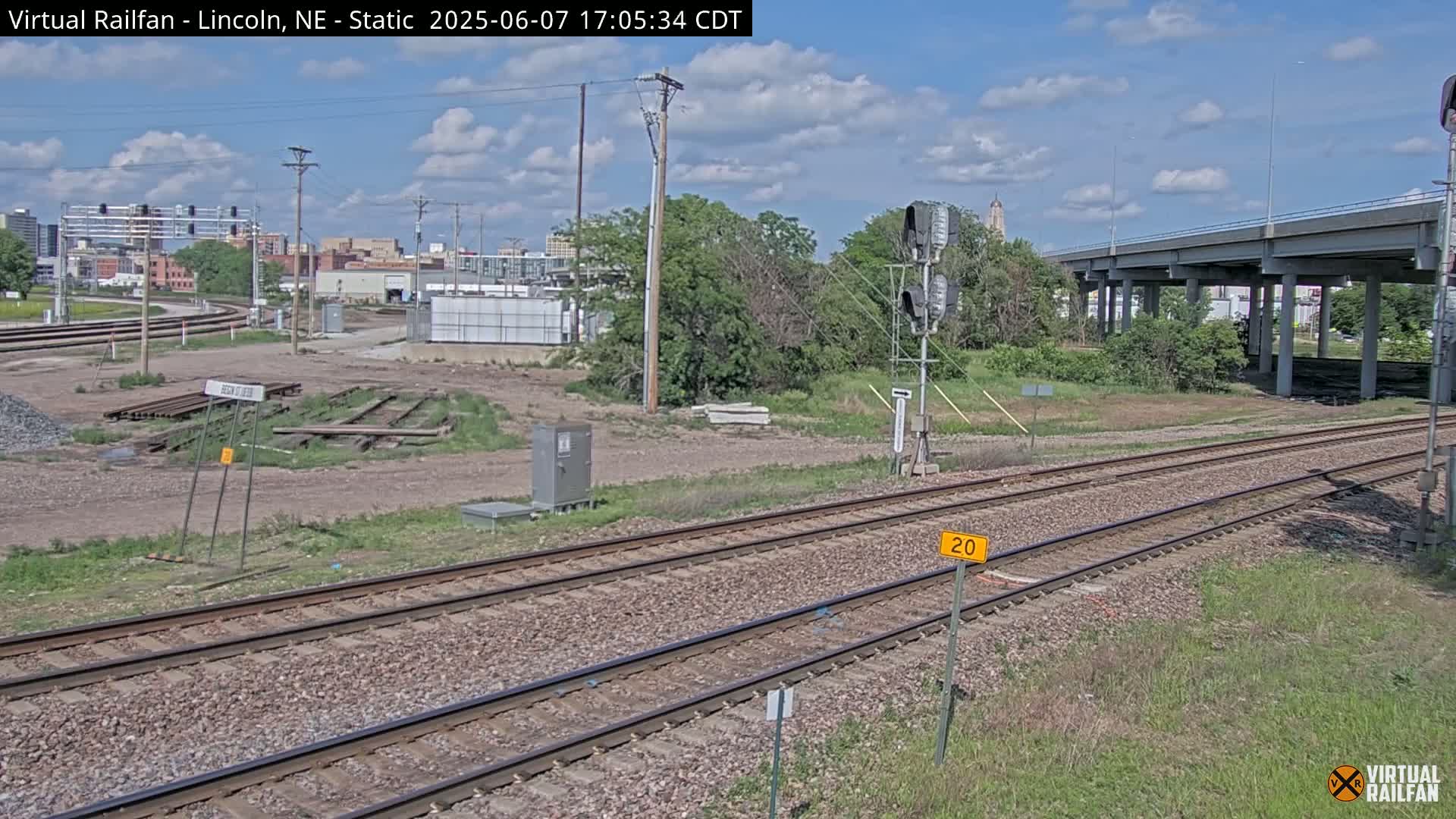 A view of railroad tracks under a partly cloudy sky, with a highway overpass in the background and some industrial buildings visible in the distance.