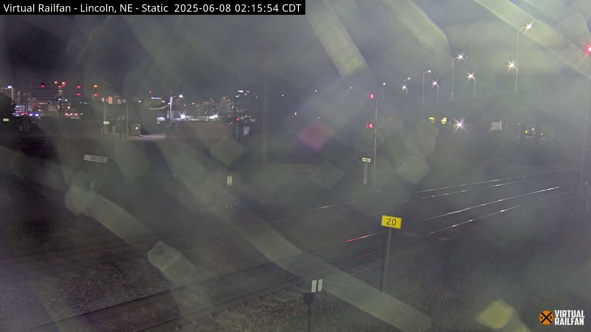 A nighttime view of railroad tracks intersecting with a road, under hazy conditions and with city lights in the background.