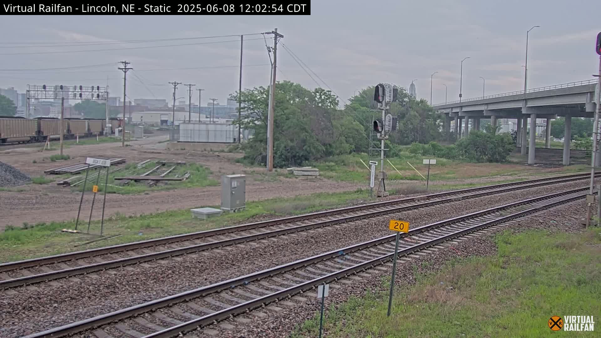 A view of railroad tracks under an overcast sky, with a highway overpass in the background and some industrial buildings visible in the distance.