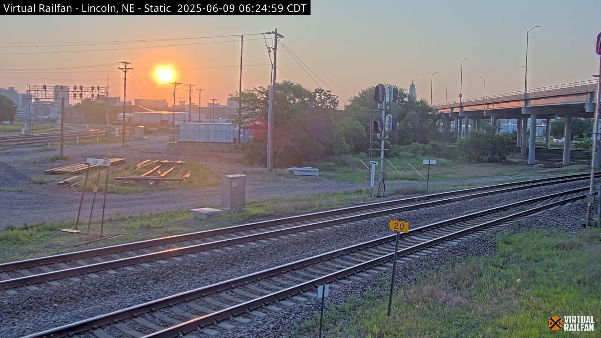 A sunrise illuminates a railroad yard with train tracks, a highway overpass, and some industrial buildings in the distance.
