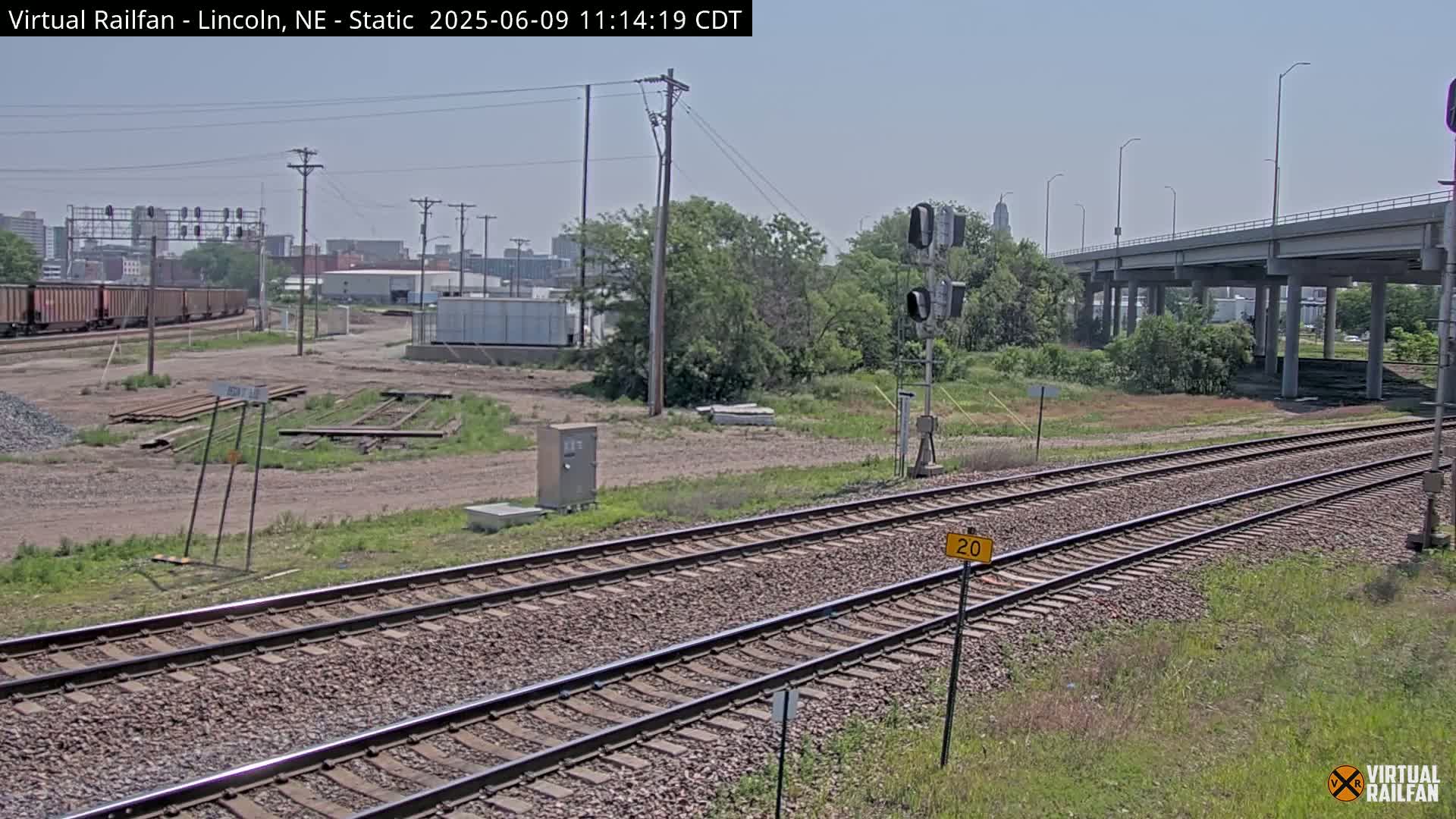A sunny day shows a railway track splitting into two, with a train in the distance, under a highway overpass and surrounded by vegetation and urban buildings in the background.