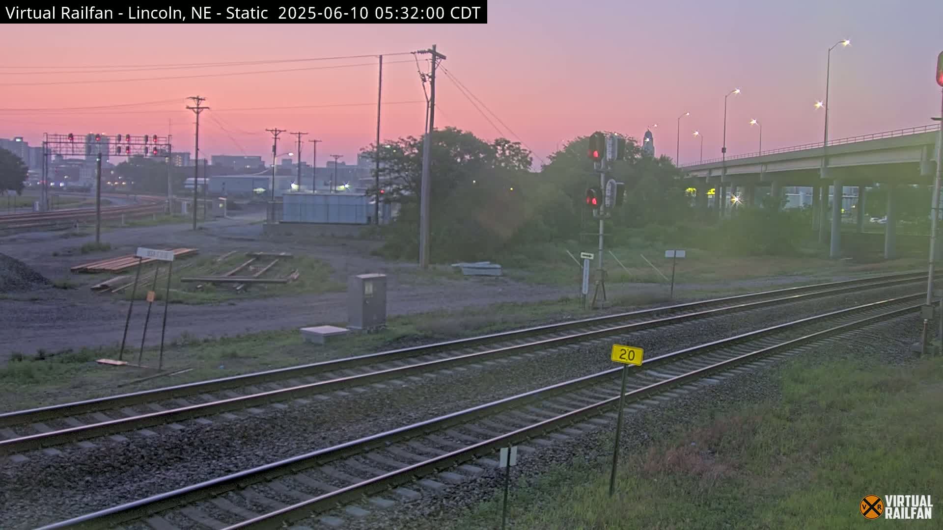 Two sets of railroad tracks run alongside a highway overpass under a pinkish sunrise.