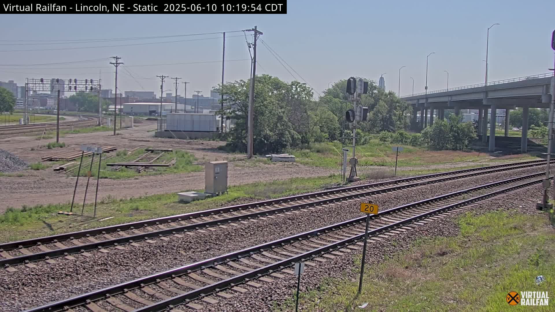 Railroad tracks extend into the distance under a clear sky, passing beneath a highway overpass with vegetation and some industrial buildings in the background.