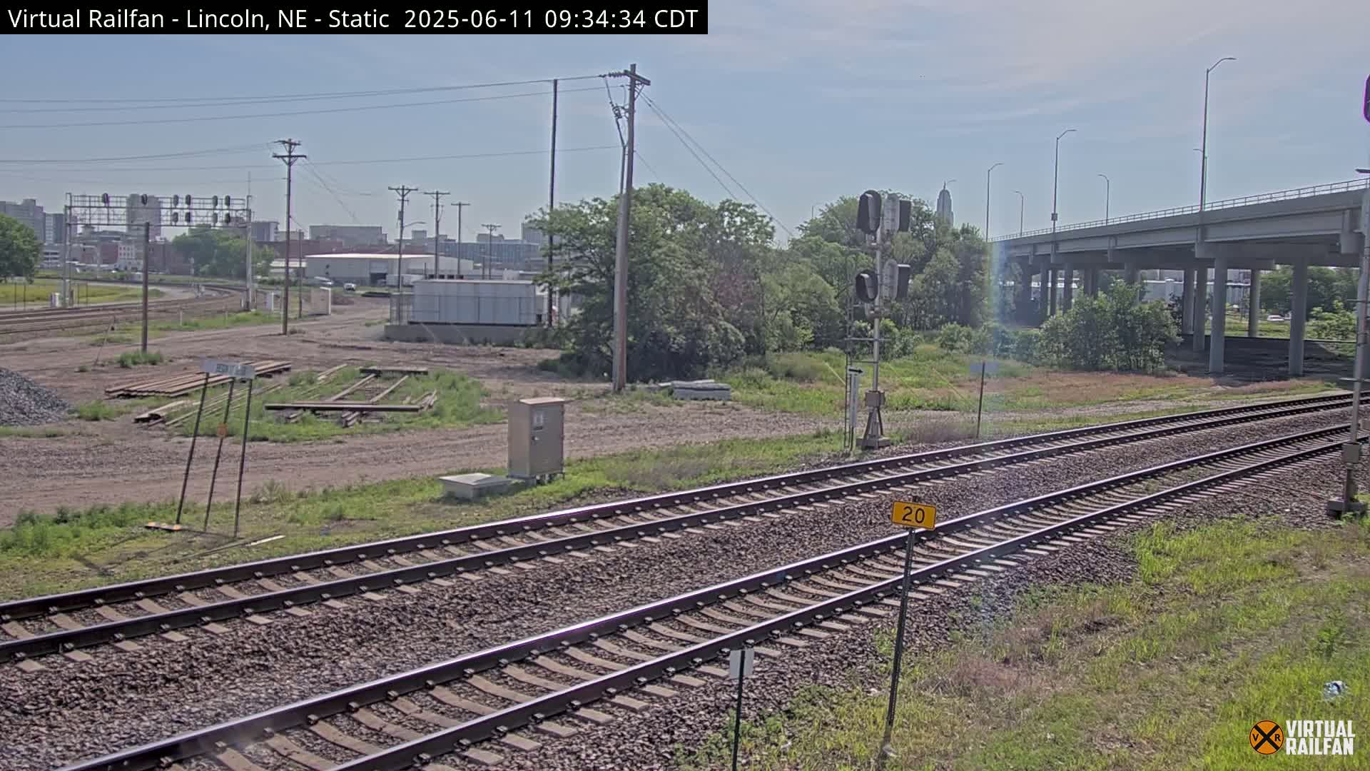 A view of railroad tracks on a partly sunny day, with a highway overpass in the background and some industrial buildings in the distance.