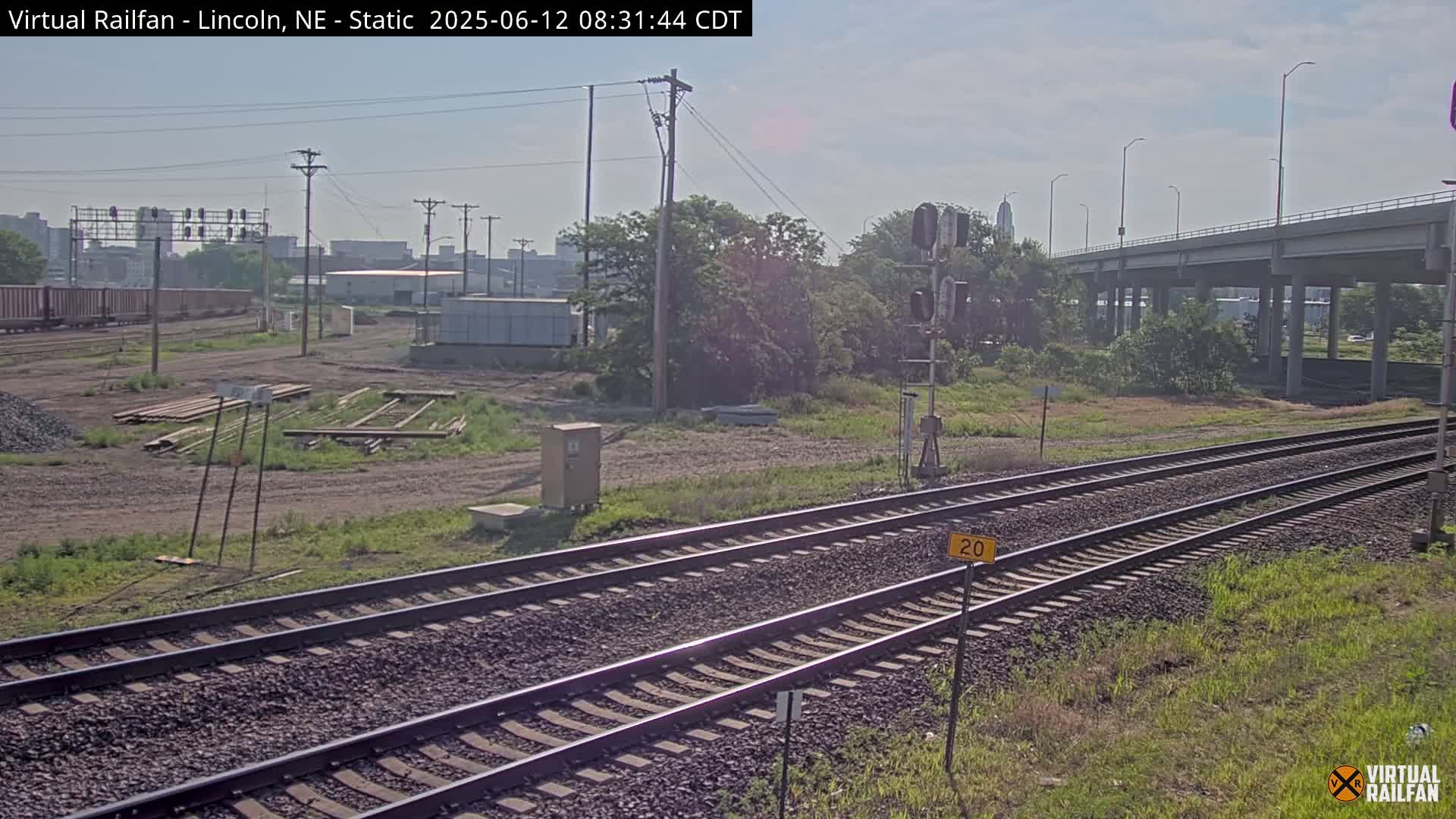 Two sets of railroad tracks extend into the distance under a mostly clear sky, passing beneath a highway overpass in a scene featuring industrial buildings and sparse vegetation.
