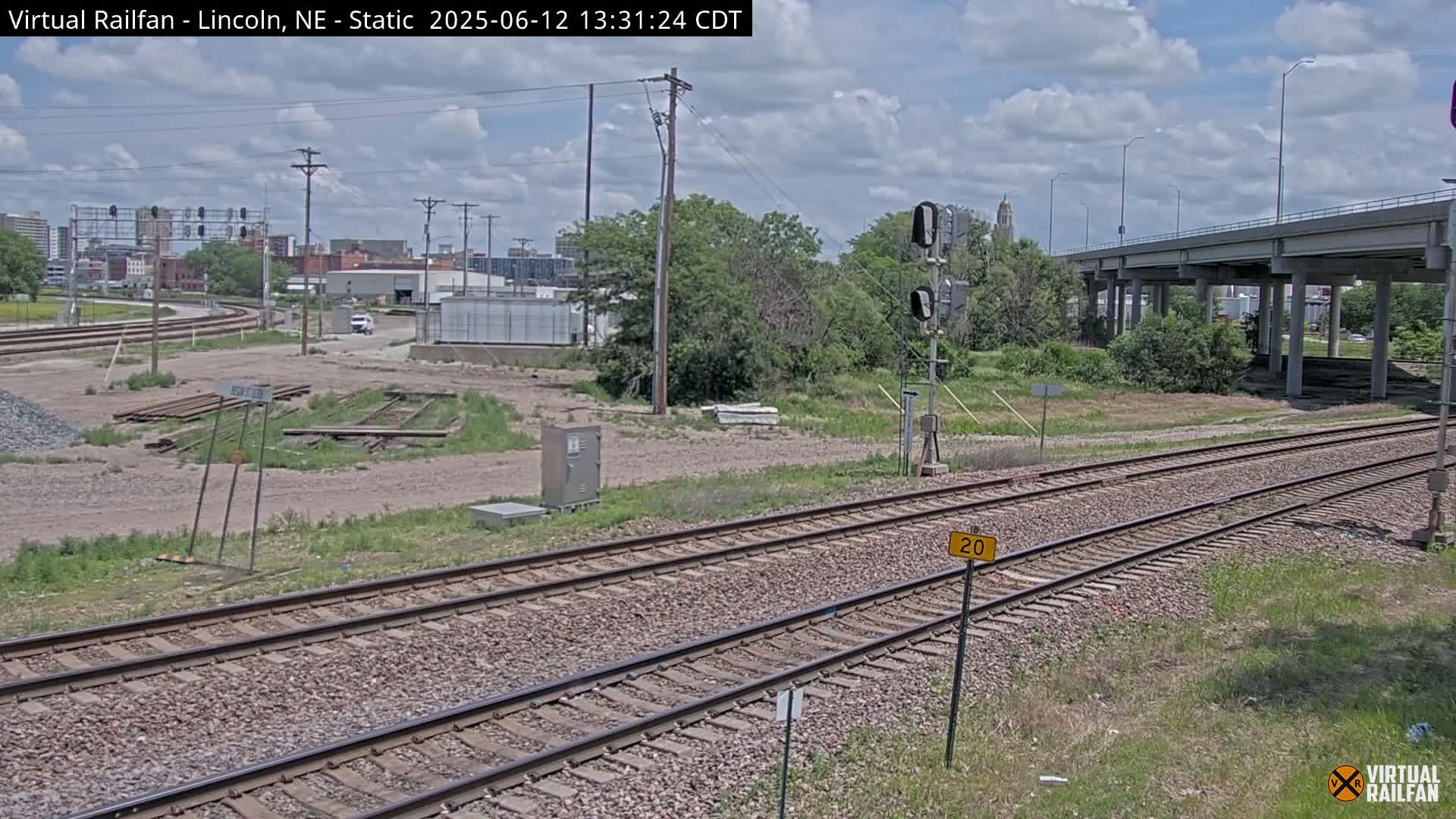 Two parallel sets of train tracks stretch into the distance under a partly cloudy sky, passing beneath a highway overpass and through a largely undeveloped area with some vegetation.