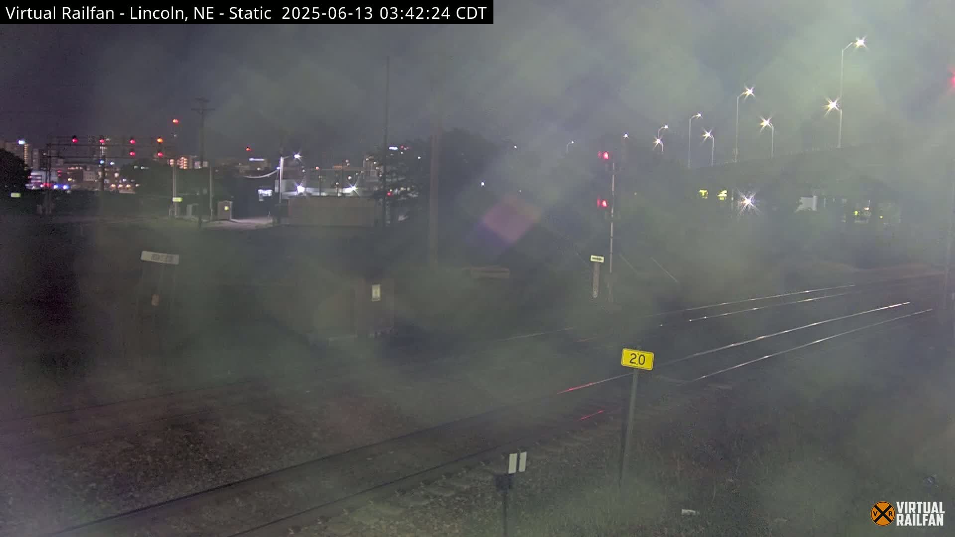 A nighttime view of railroad tracks, a street, and a hazy cityscape in the distance under a foggy sky.