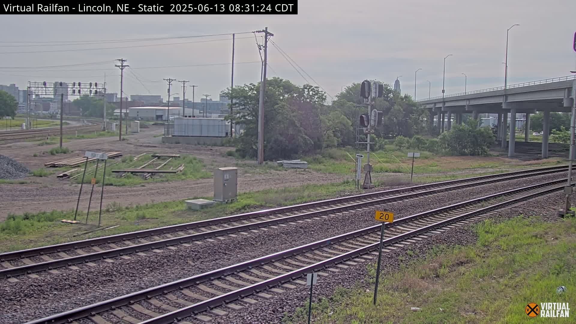 Two sets of railroad tracks extend into the distance under an overcast sky, passing beneath a highway overpass and alongside some undeveloped land with scattered vegetation.