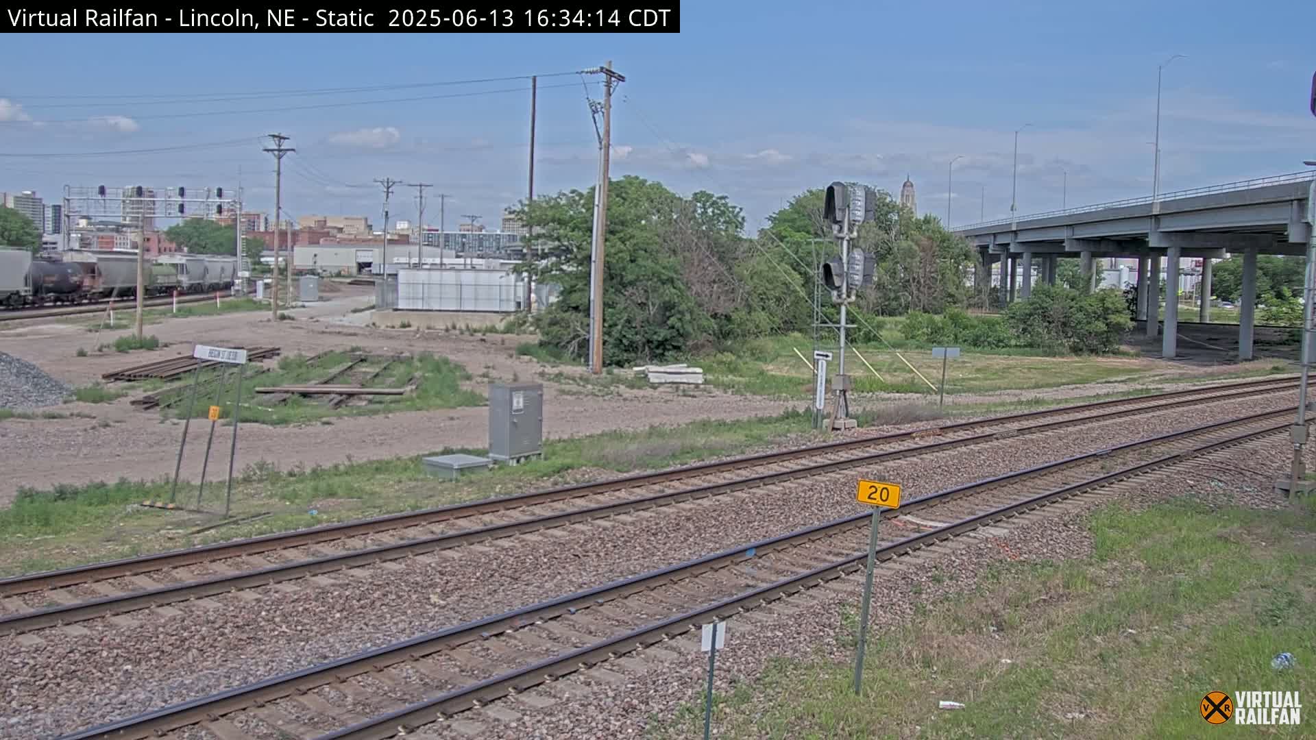 Under a partly cloudy sky, a view of railroad tracks stretches towards a highway overpass and a city skyline in the distance, with some industrial buildings and sparse vegetation surrounding the tracks.
