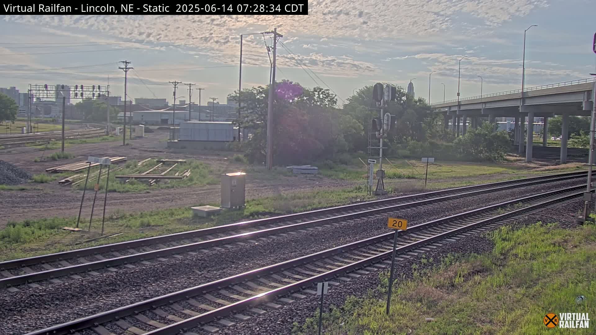A partly cloudy sky overlooks railroad tracks, a highway overpass, and a mostly barren landscape with some vegetation and industrial buildings in the distance.