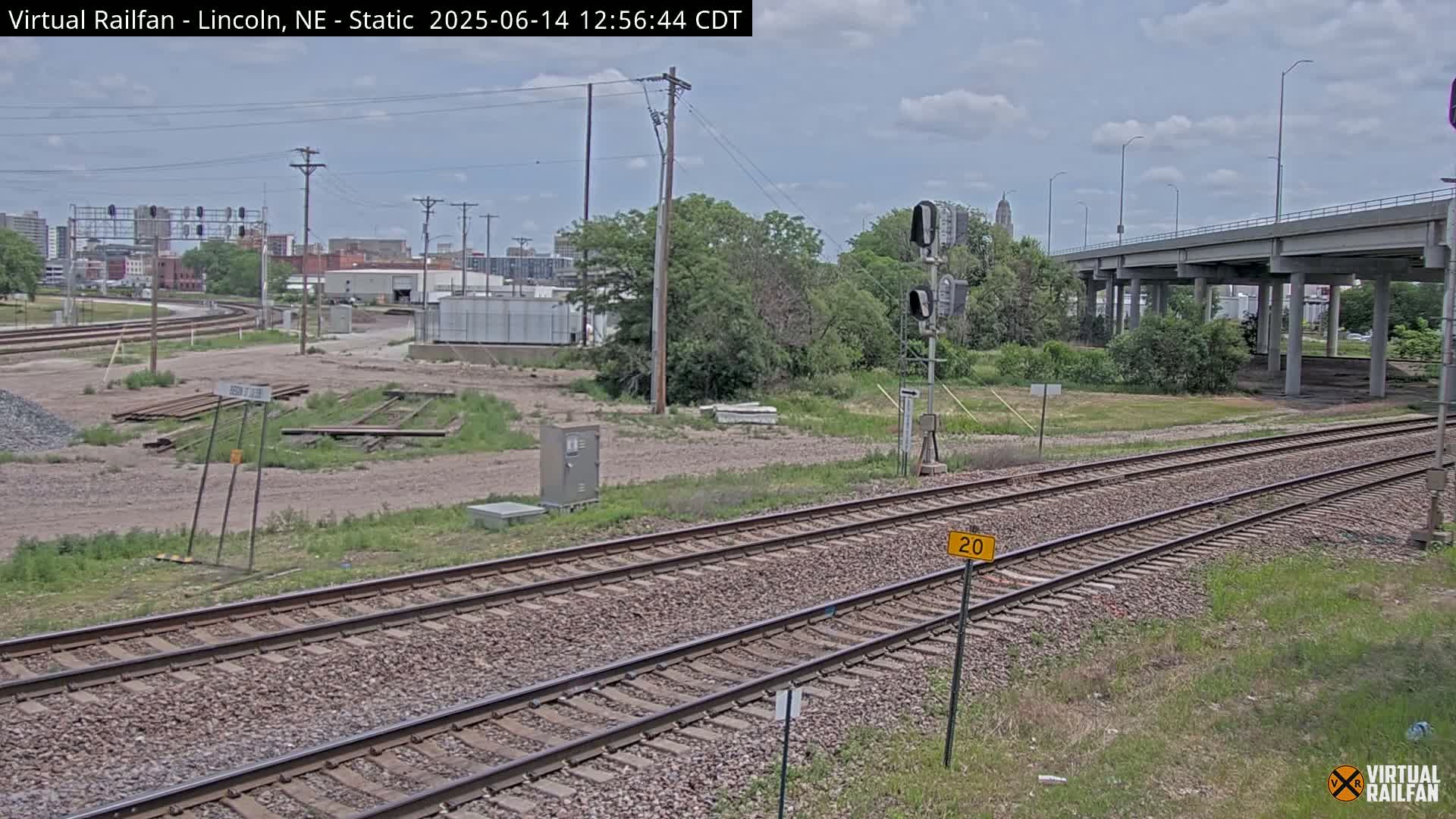 A view of railroad tracks on a partly cloudy day, with a highway overpass in the background and some industrial buildings visible in the distance.