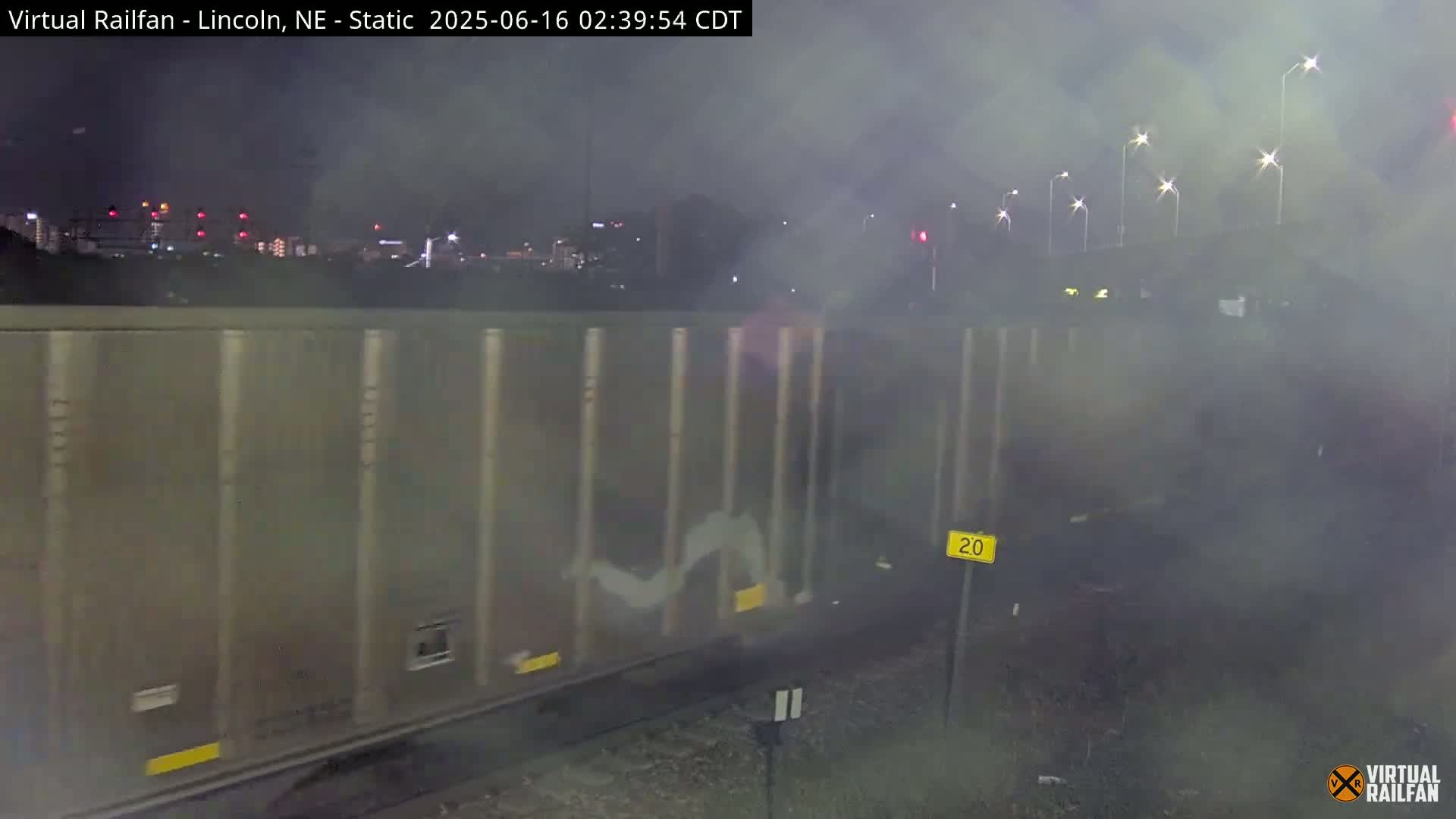 A long freight train car passes by at night under hazy conditions, with city lights visible in the background.