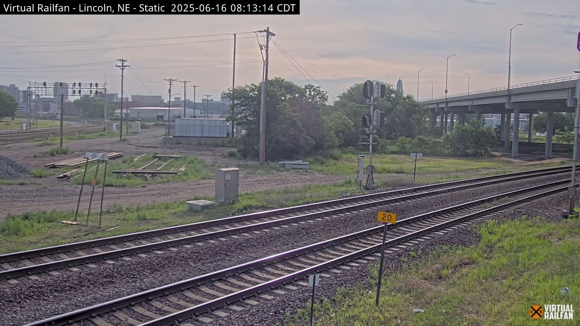 A view of railroad tracks on a partly cloudy day, with vegetation and an elevated roadway visible in the background.