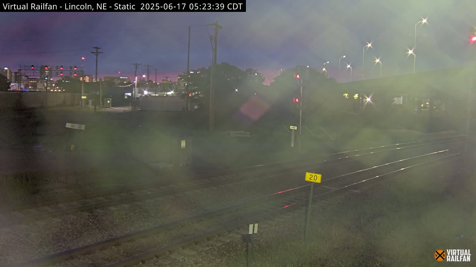 A nighttime view of railroad tracks, with a city skyline visible in the distance under a dusky sky.