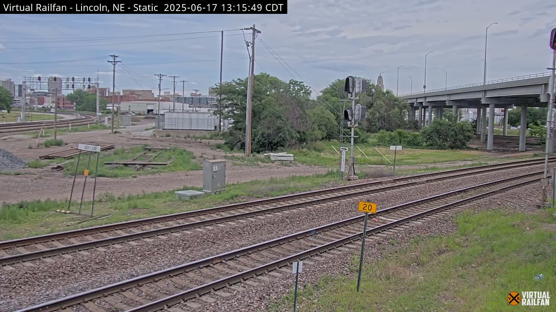 Two parallel sets of train tracks stretch towards a large overpass under an overcast sky, with sparse vegetation and industrial buildings in the background.