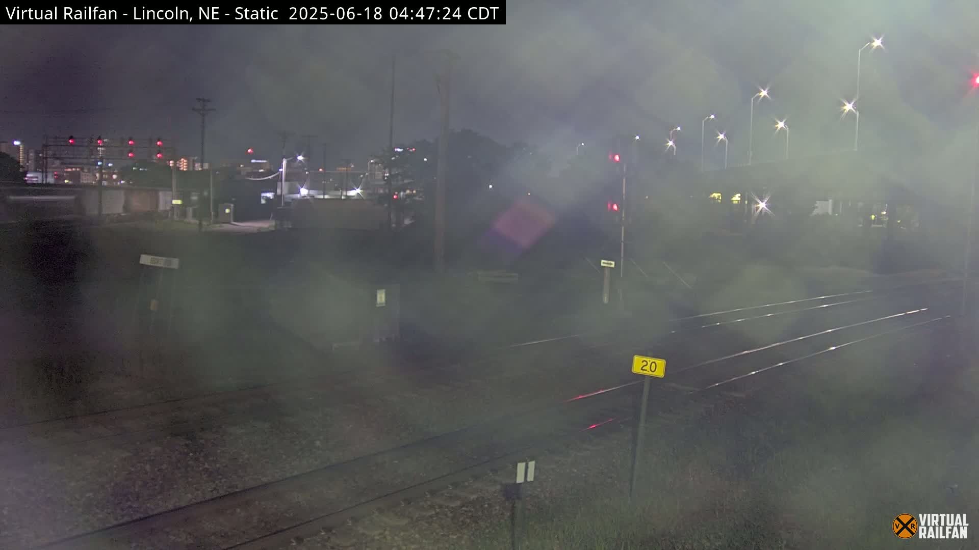 A nighttime view of railroad tracks, illuminated by streetlights and hazy with fog or mist, shows a city skyline in the background.