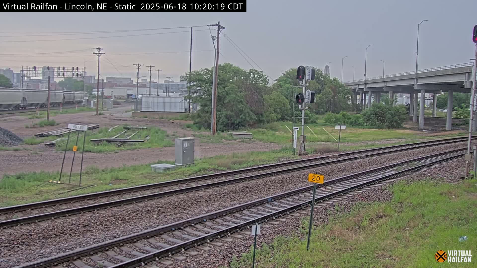 A view of railroad tracks under an overcast sky, with a highway overpass in the background and a freight train visible in the distance.