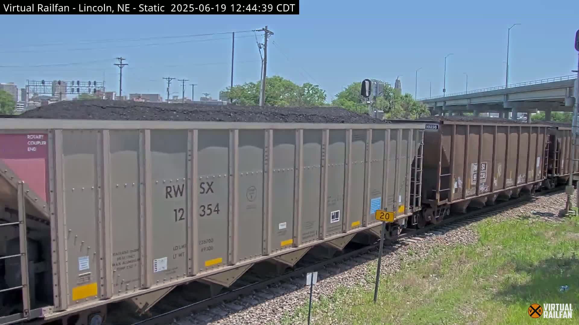 A long freight train carrying coal is shown on a sunny day, passing under a highway overpass.