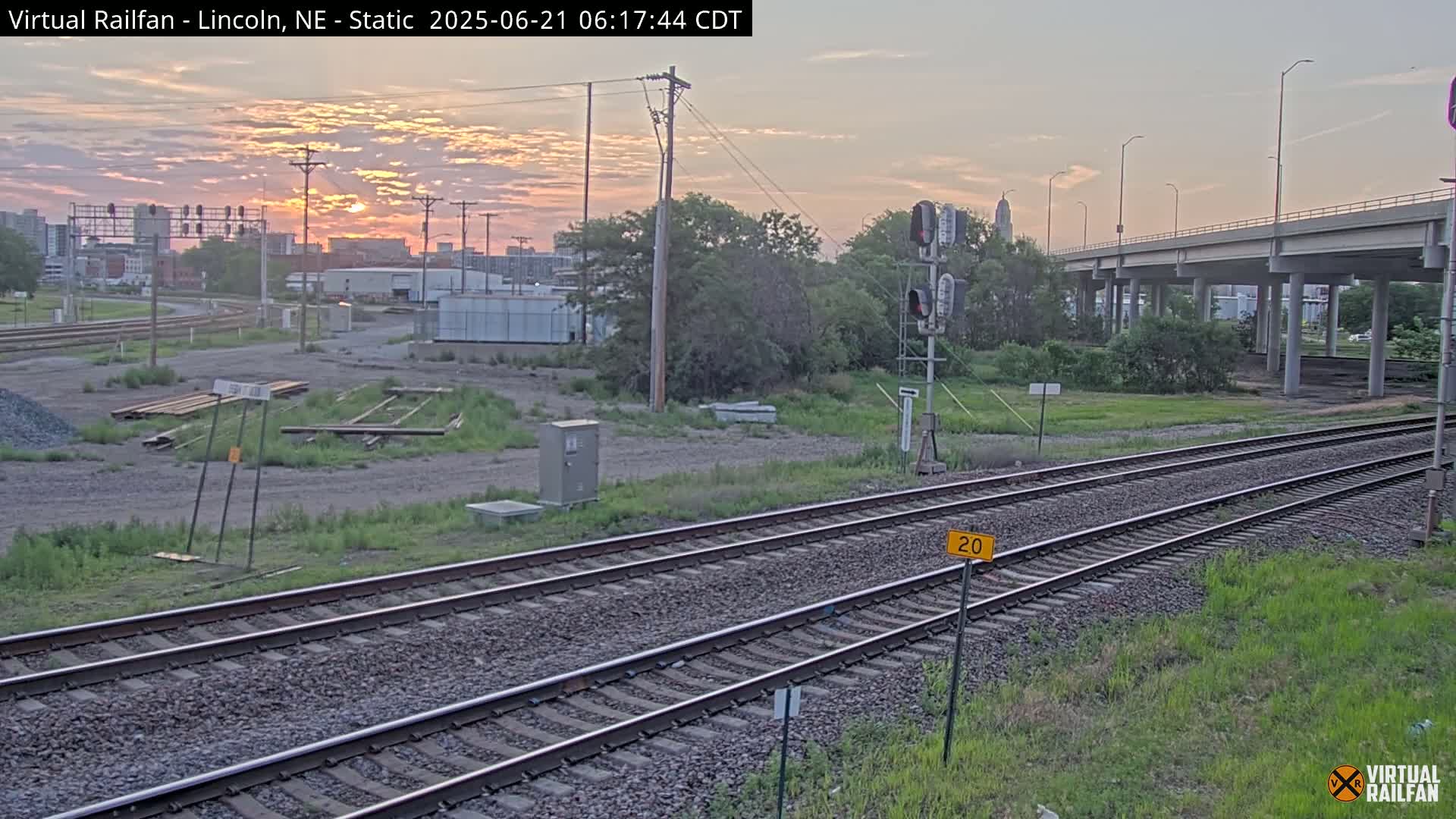 A sunrise illuminates a railroad track and surrounding industrial area, under a mostly clear sky.
