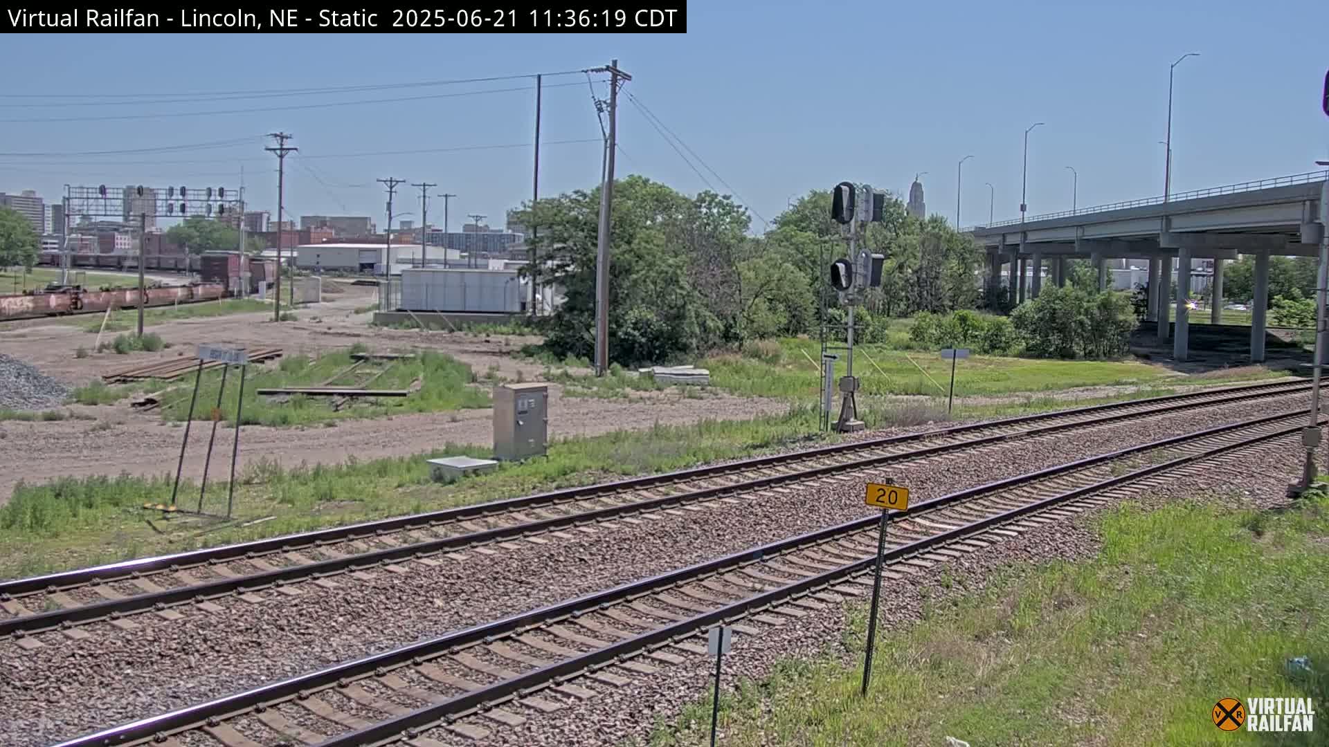 Under a clear sky, two sets of train tracks extend from the foreground toward a city skyline and a large overpass, with some vegetation and a few industrial buildings in between.
