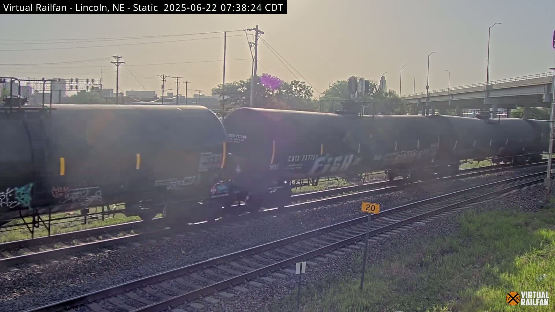 A long freight train of dark-colored tanker cars sits on railroad tracks under a hazy, sunny sky.