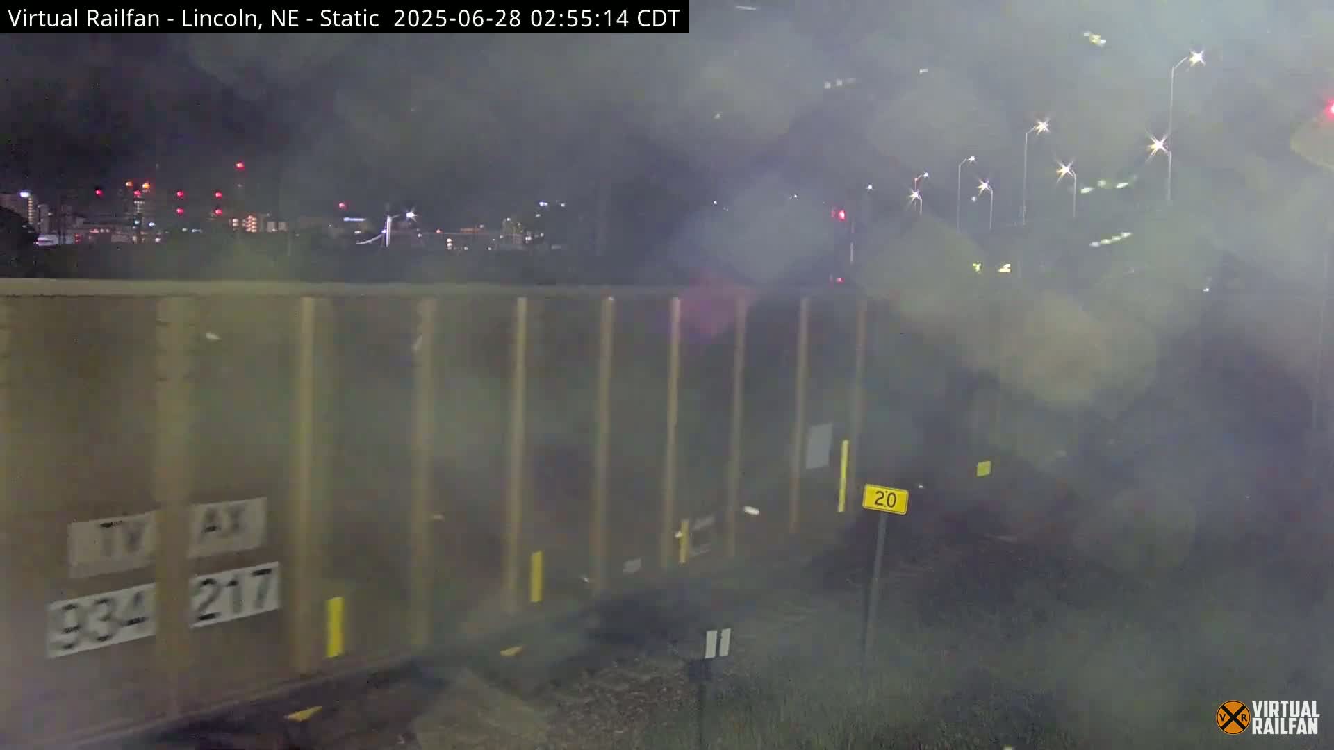 A tan freight train car is partially visible at night in foggy conditions, with a city skyline in the background.