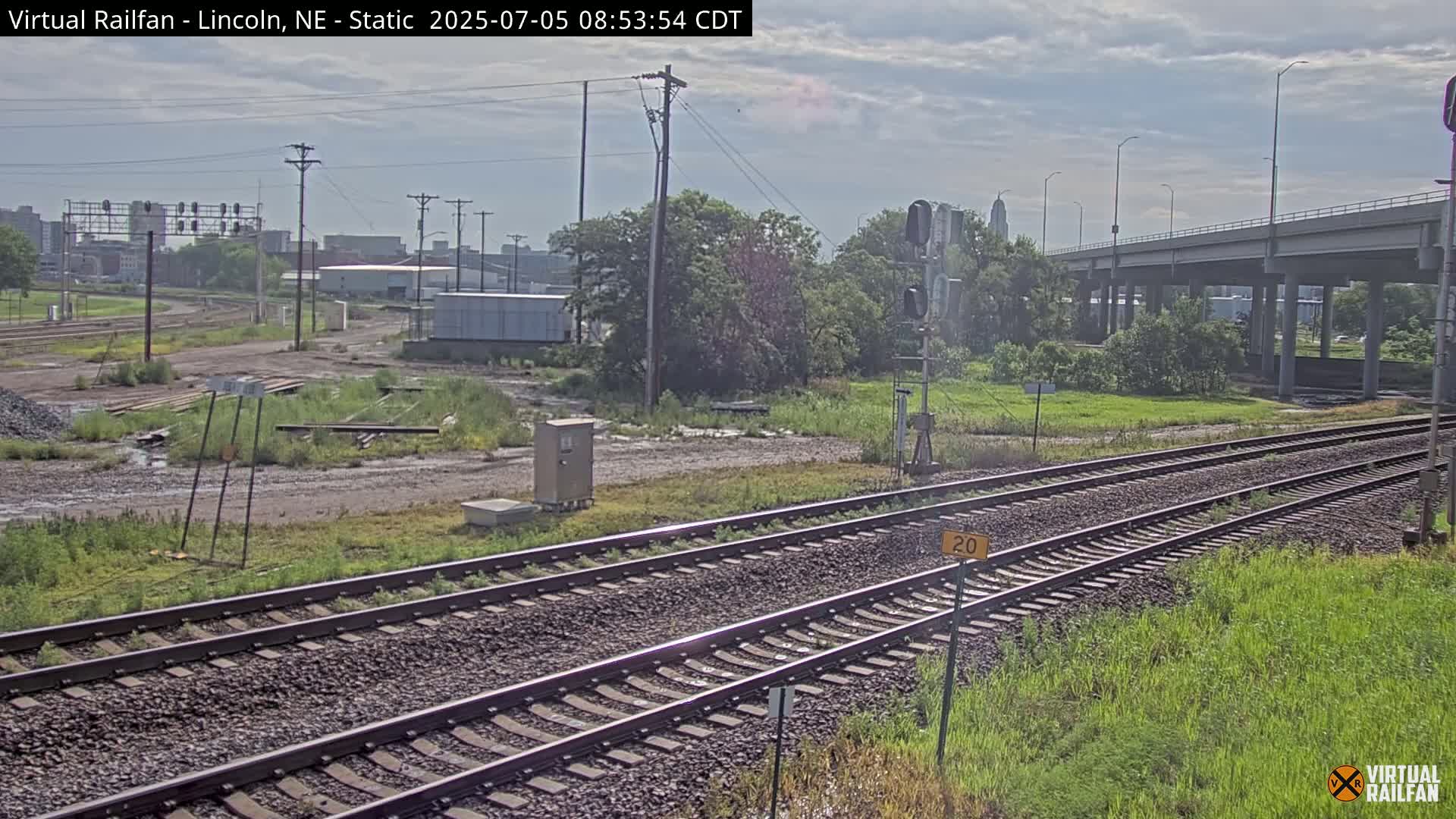 Two sets of railroad tracks stretch into the distance under a partly cloudy sky, passing under a highway overpass and bordered by patchy grass and weeds.