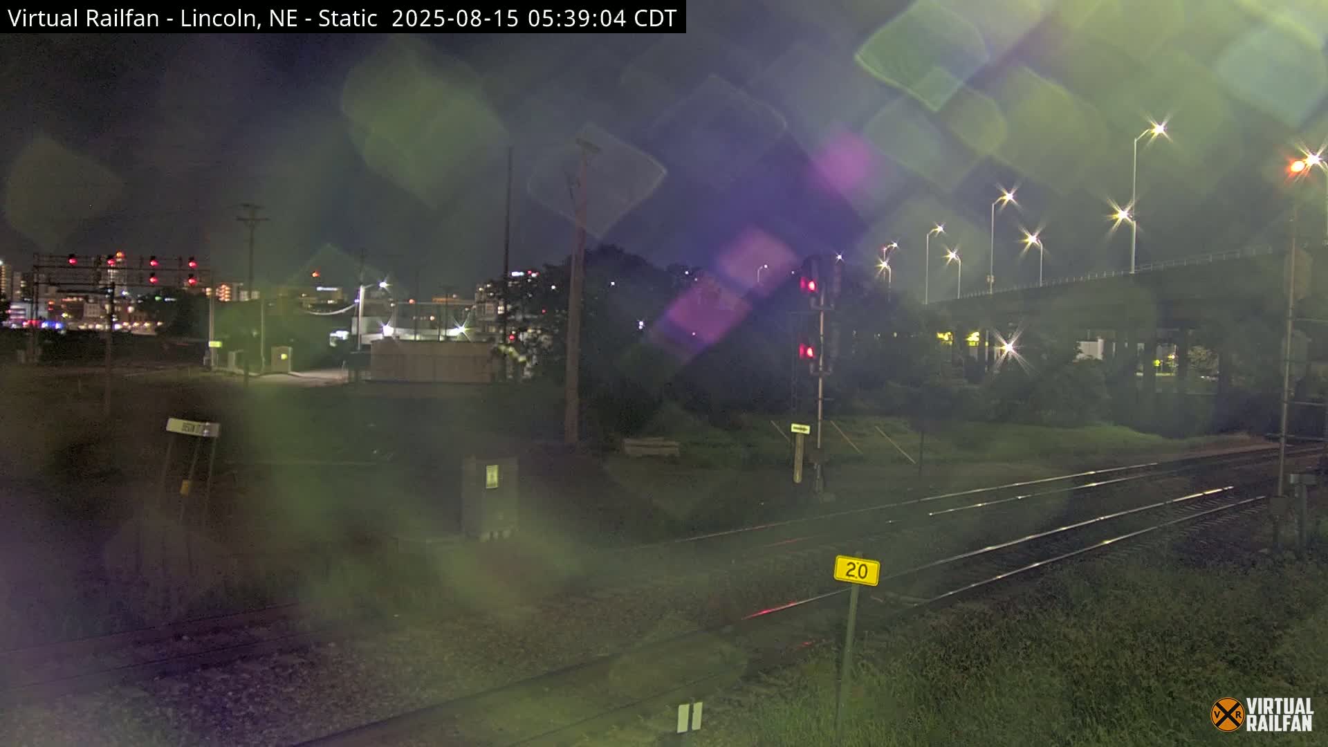 A nighttime view of railroad tracks under a mostly clear sky, with lights illuminating a nearby overpass and cityscape in the distance.