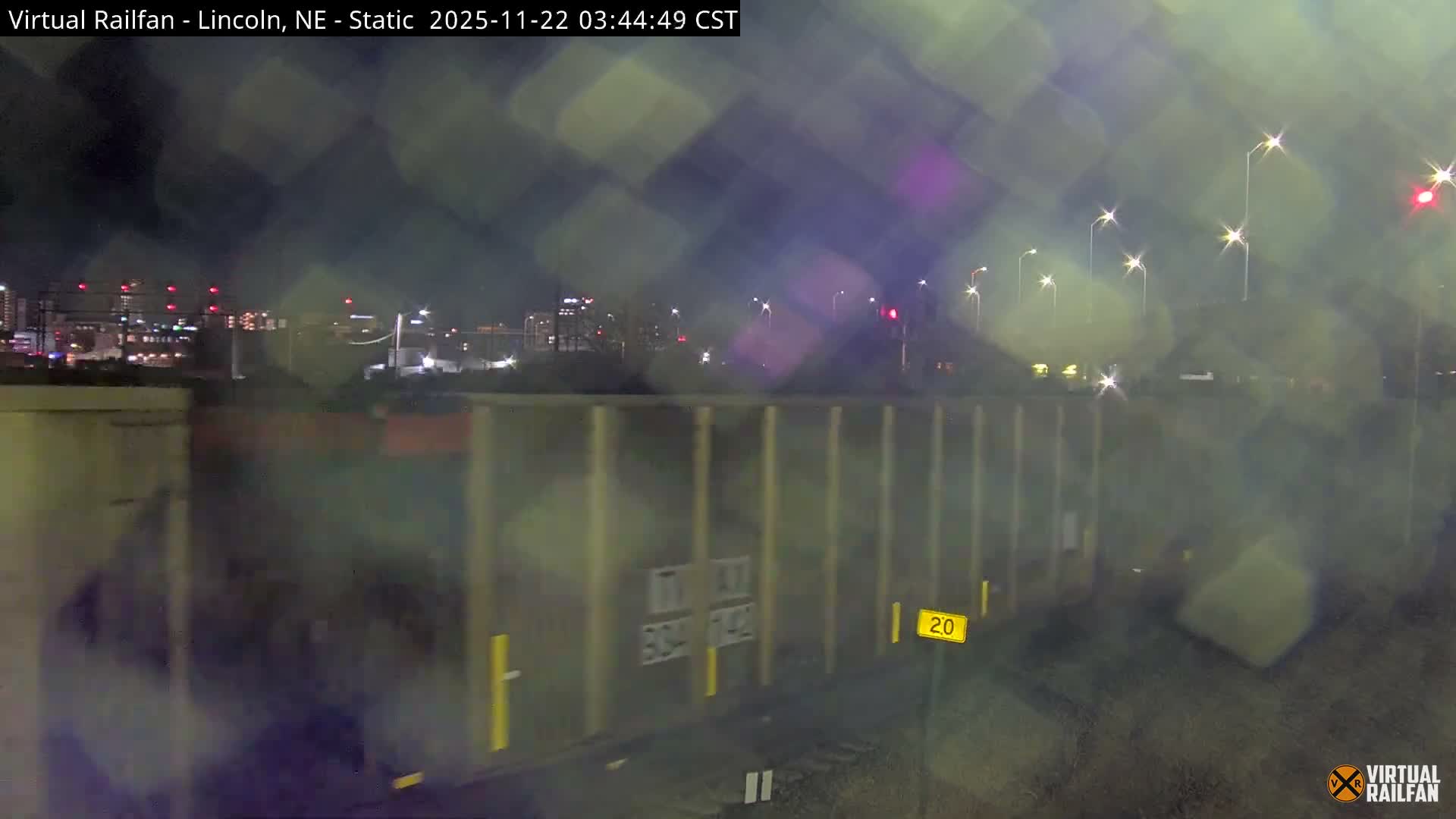 A freight train passes through an urban landscape at night under rainy or very damp conditions, causing the visible lights to appear diffused and creating starburst effects across the image.