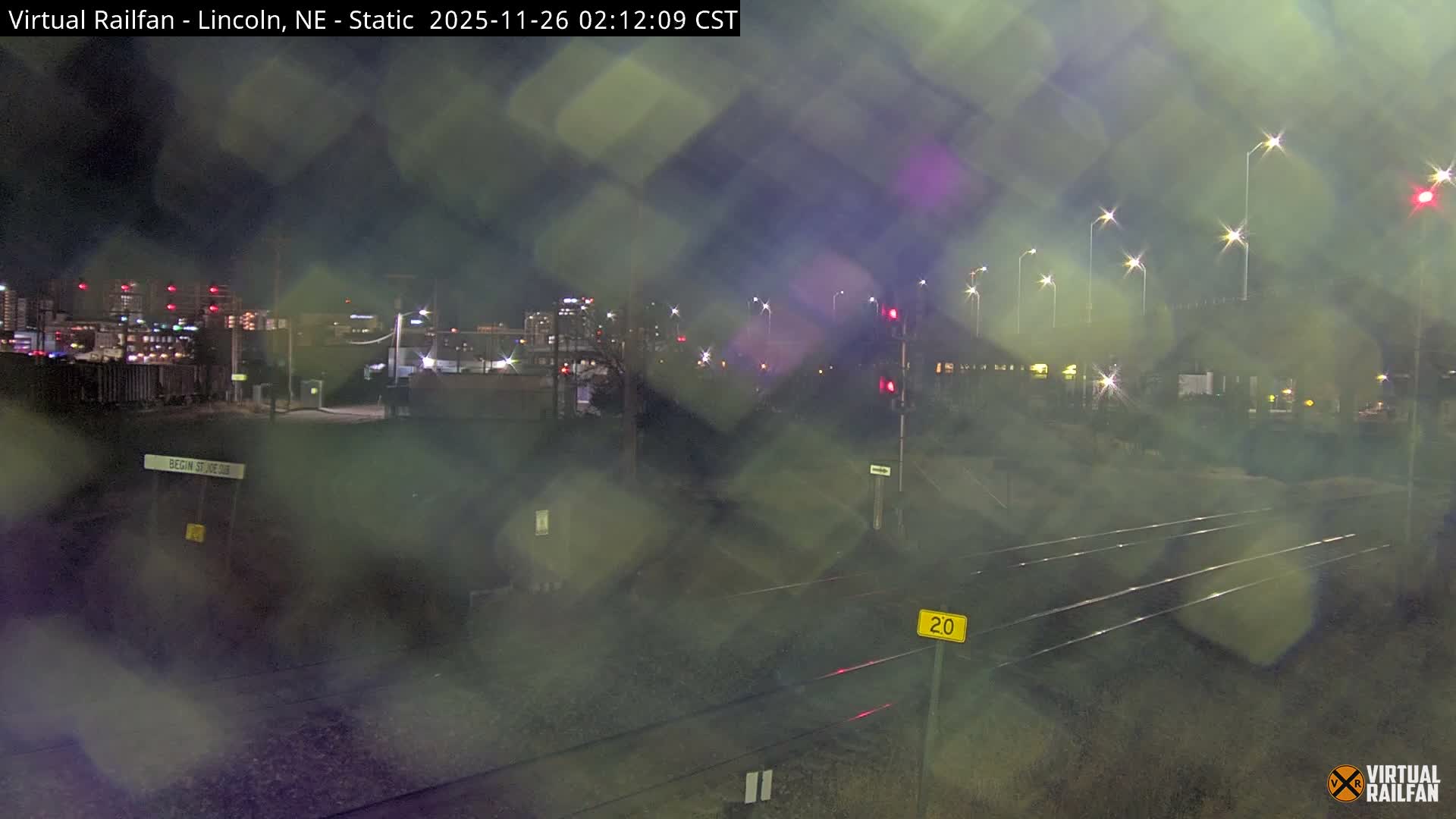A rainy or misty urban rail yard at night, viewed through a wet camera lens, features multiple tracks stretching into the distance alongside brightly flaring streetlights, red traffic signals, and the illuminated skyline of a distant city.