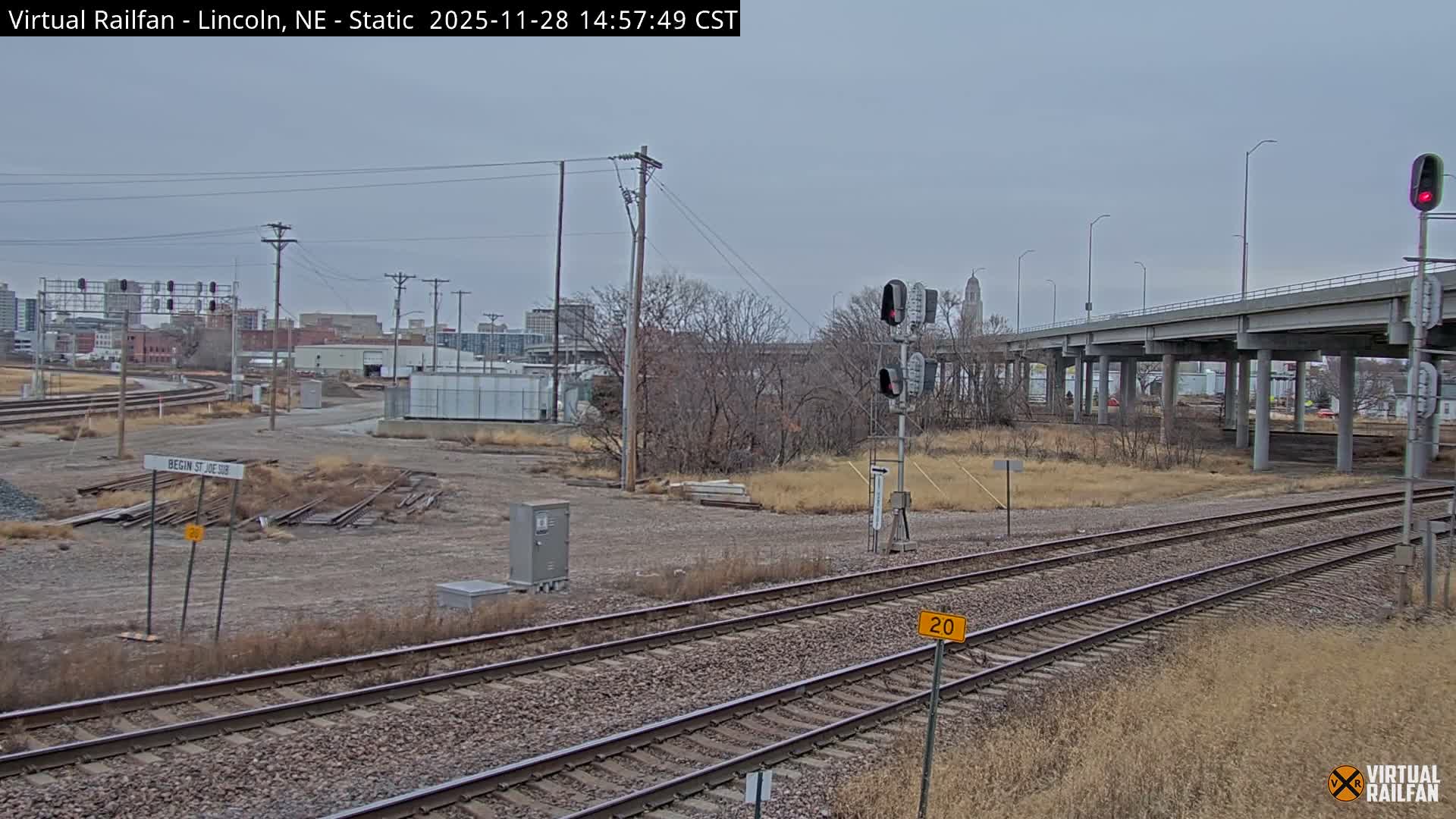 Numerous railroad tracks curve through a dry, late-season urban landscape featuring an elevated highway overpass, distant city buildings, and various railroad signals, all under a uniformly cloudy and overcast sky.