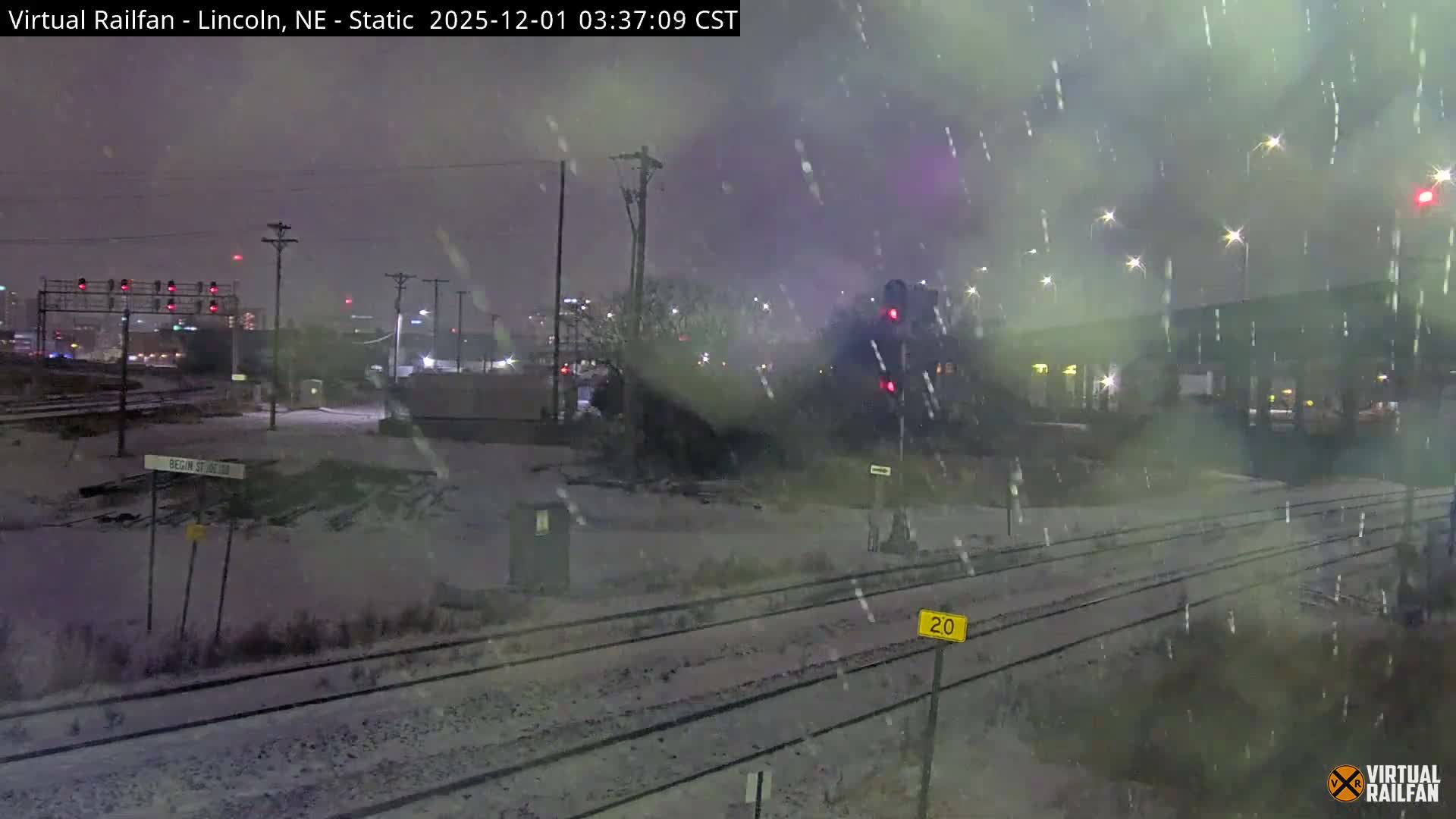 The image depicts a snowy urban night scene looking over multiple railroad tracks covered in fresh snow, with various red traffic signals, streetlights, and distant city lights illuminating the falling flakes.