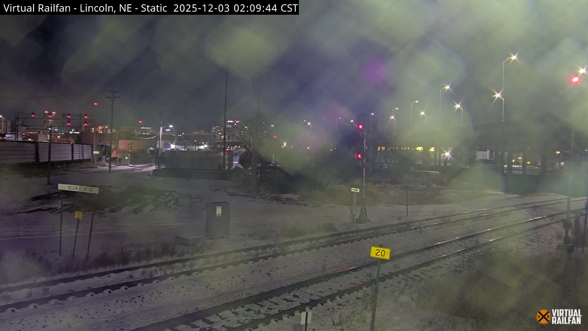A nighttime urban landscape features snow-covered railroad tracks stretching into the distance, illuminated by glowing streetlights and city lights under hazy or misty conditions.