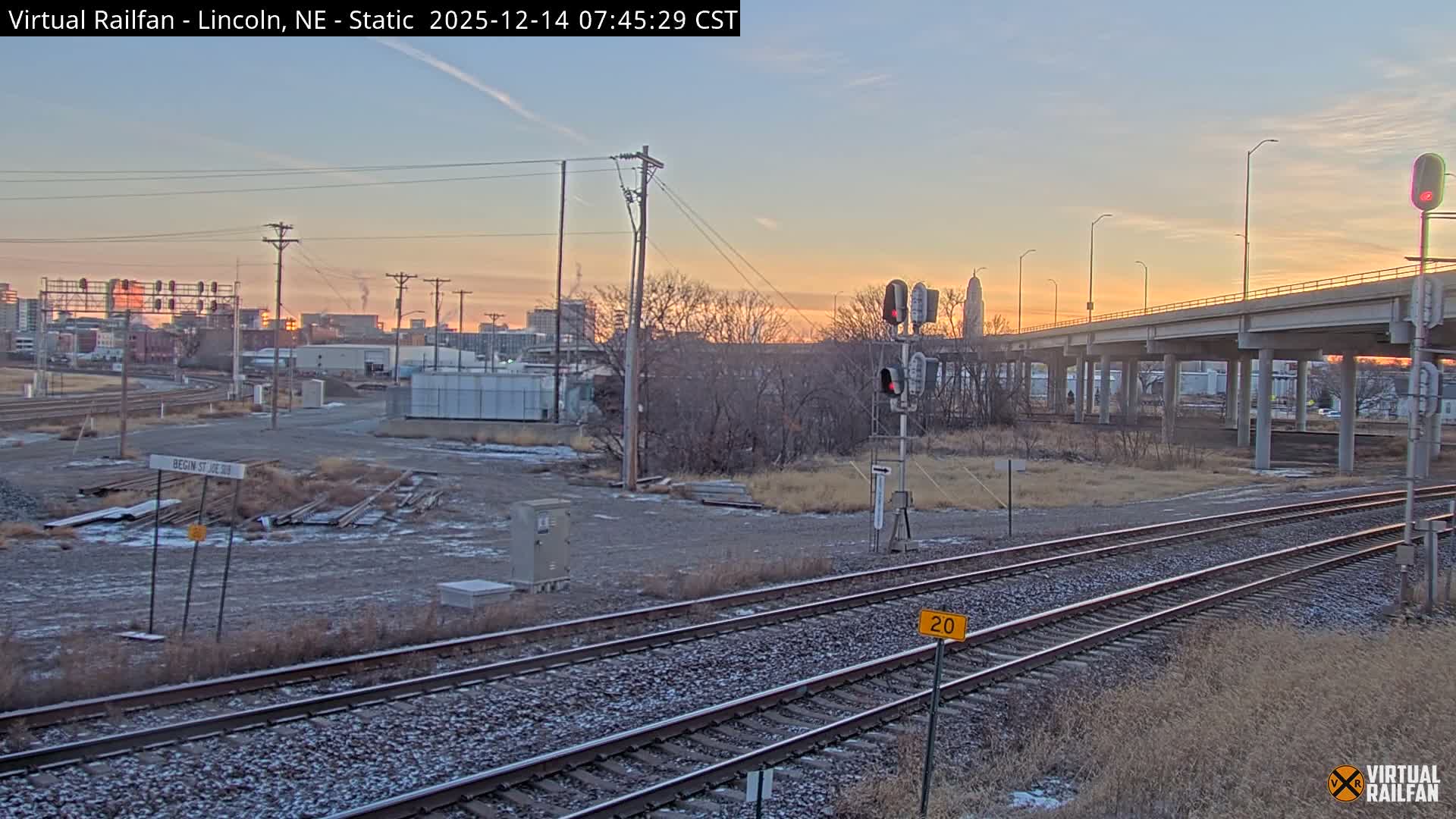 A nighttime urban landscape features snow-covered railroad tracks stretching into the distance, illuminated by glowing streetlights and city lights under hazy or misty conditions.