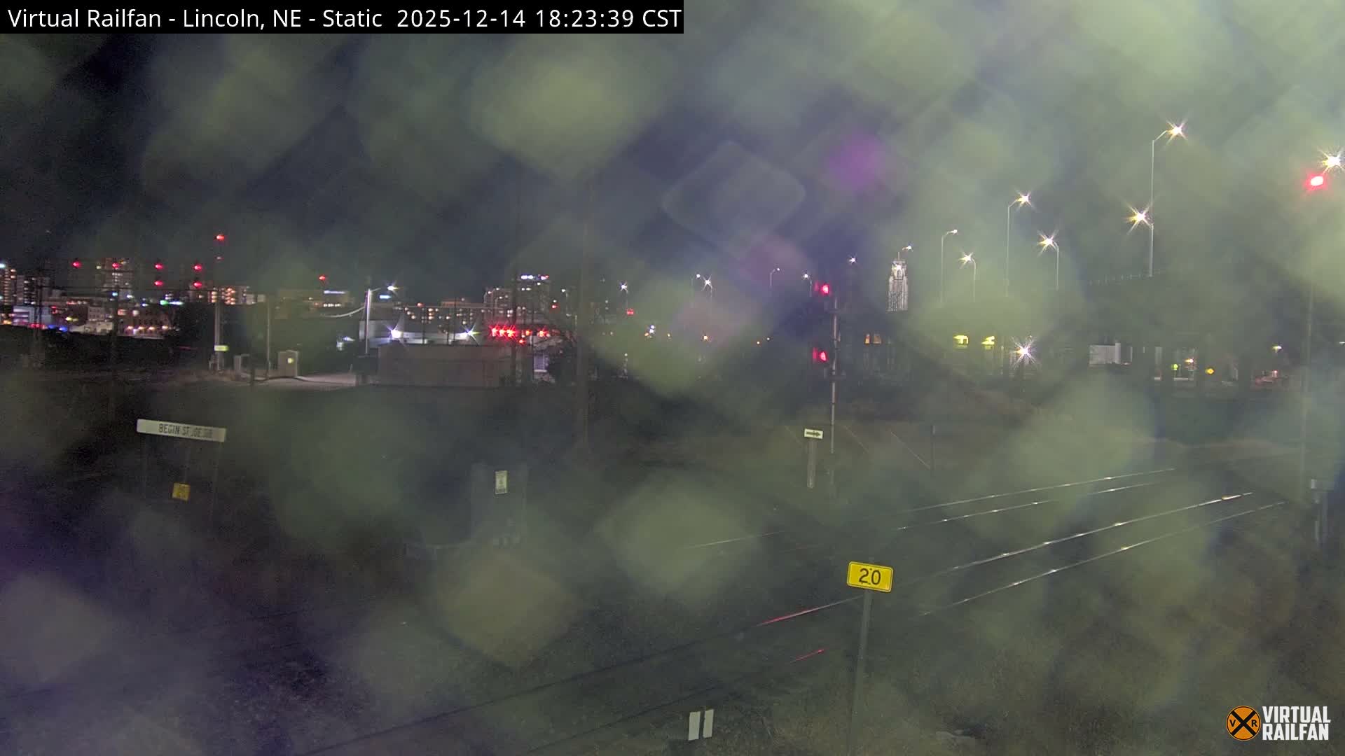 A nighttime urban landscape features snow-covered railroad tracks stretching into the distance, illuminated by glowing streetlights and city lights under hazy or misty conditions.