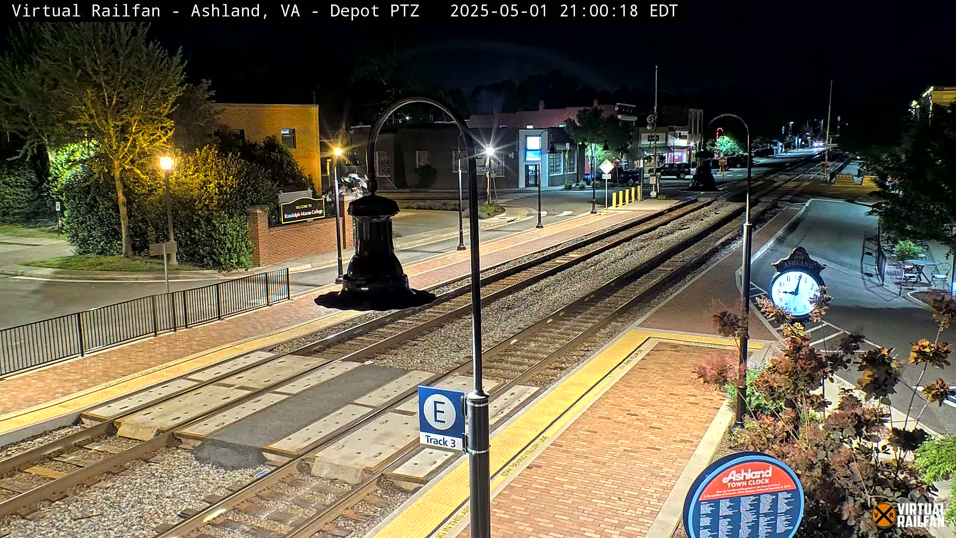 At night, a train station platform with tracks and brick pavement is illuminated by streetlights, showing some buildings and landscaping in the background.