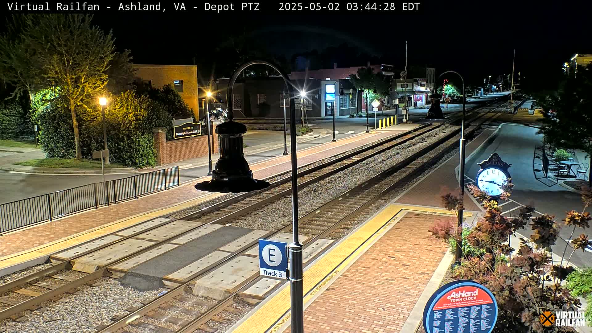 A nighttime view of a train station platform with tracks, brick pavement, streetlights, and landscaping.