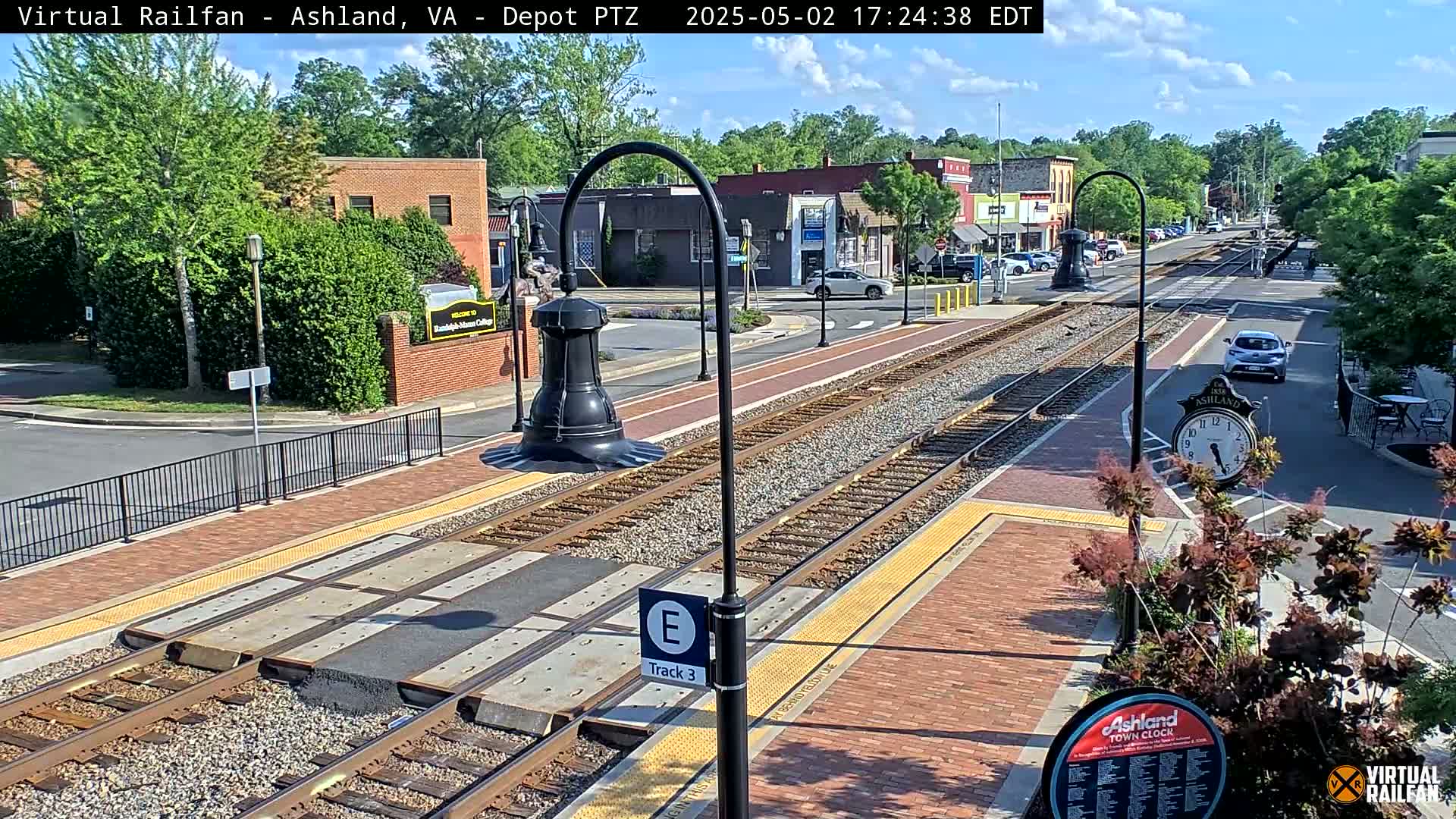A sunny day view of a train station platform with tracks, brick pavement, and surrounding buildings and trees.