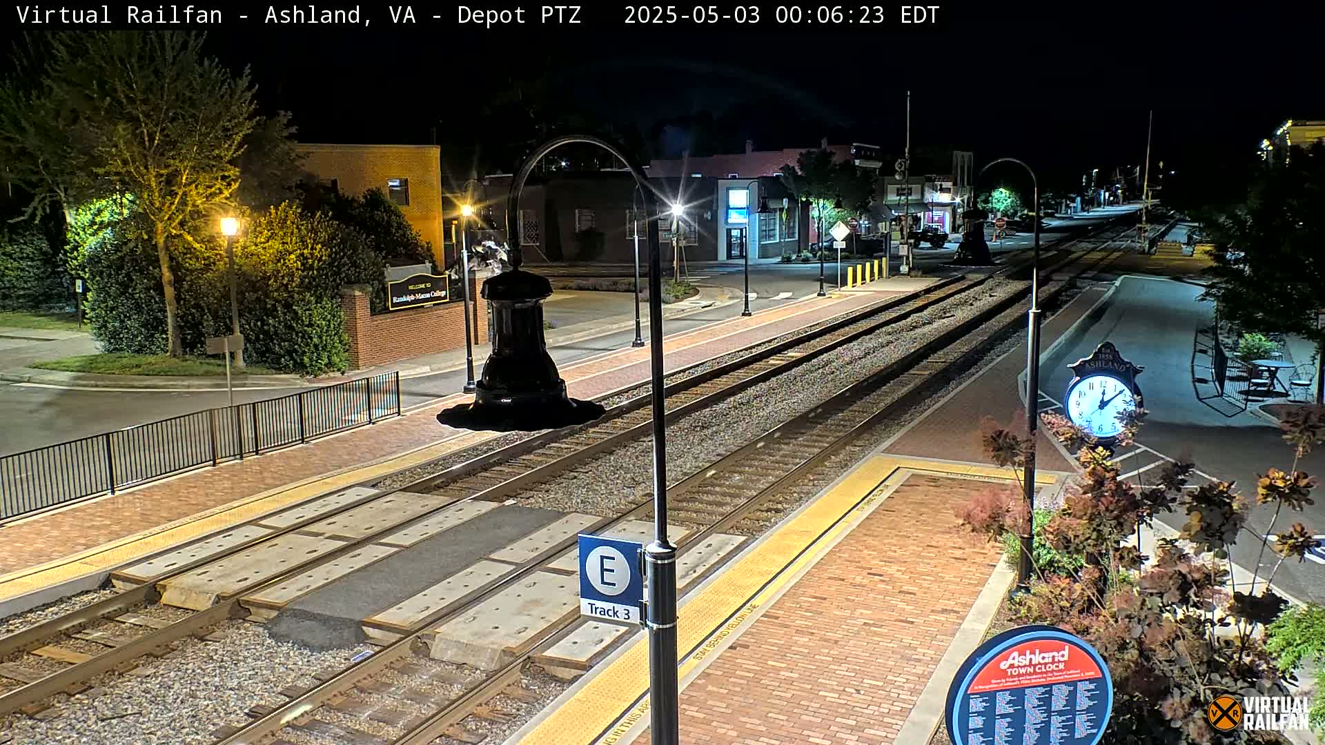 A nighttime view of a train station platform with train tracks, brick pavement, and streetlights, under clear skies.