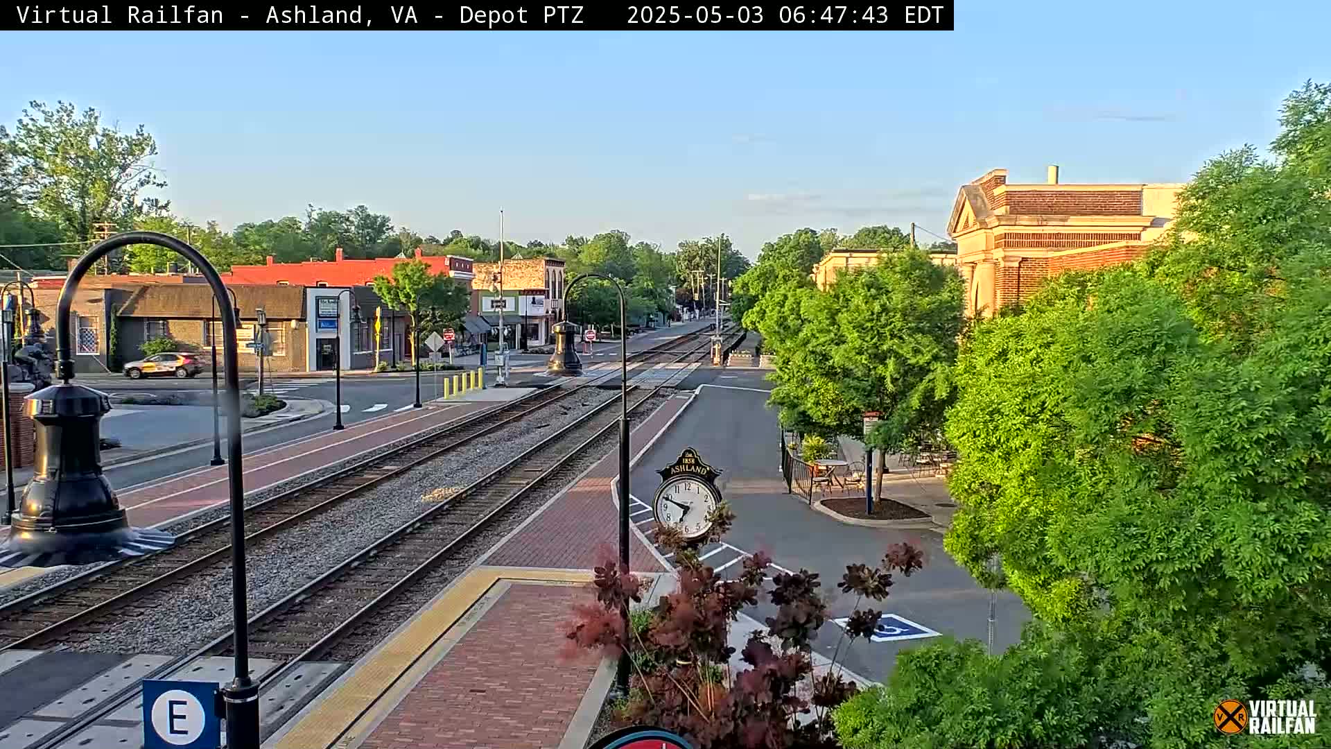 A train track runs through a town square on a clear, sunny morning, with leafy green trees lining the streets and brick buildings in the background.